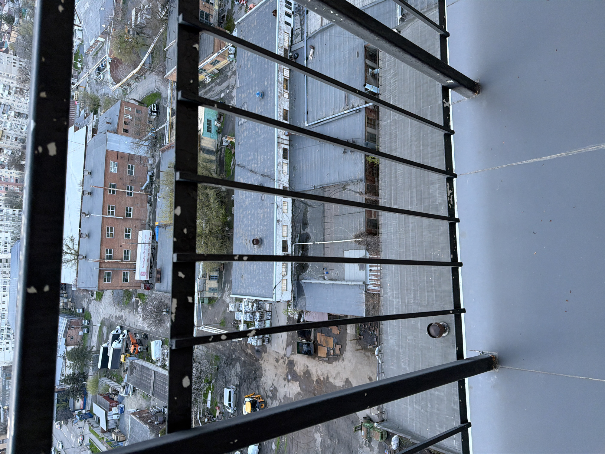 View from a high balcony in Rostov-on-Don, Russia, through a black metal railing. The view shows a cityscape with various buildings, including residential apartments and industrial structures with gray roofs. Construction or industrial activity is visible in a yard below, featuring machinery, piles of materials, and vehicles. The sky is overcast, indicating a cloudy day.