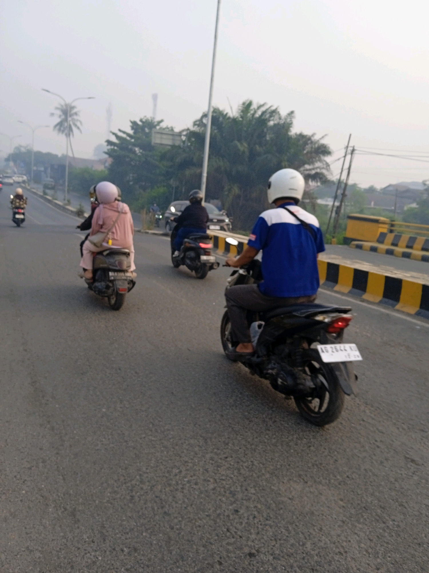 An eye-level, medium shot captures a hazy street scene in Banjarmasin, Indonesia, with several motorcycles on an asphalt road. In the foreground, a person wearing a white helmet and a blue and white shirt rides a black motorcycle. Their legs are clad in grey trousers, and they are wearing sandals. The license plate of this motorcycle reads "AG 2644 KL". To the left, another motorcycle is visible with two occupants. The rider wears a dark helmet, while the passenger in front wears a pink helmet and a pink jacket. A beige bag is slung across the passenger's shoulder. Further down the road, another motorcycle with a rider wearing a black helmet is seen. In the background, palm trees and other vegetation are present, with power lines and poles in the distance. A yellow and black striped barrier runs along the right side of the road, indicating a bridge or overpass. The sky is overcast, contributing to the hazy atmosphere.