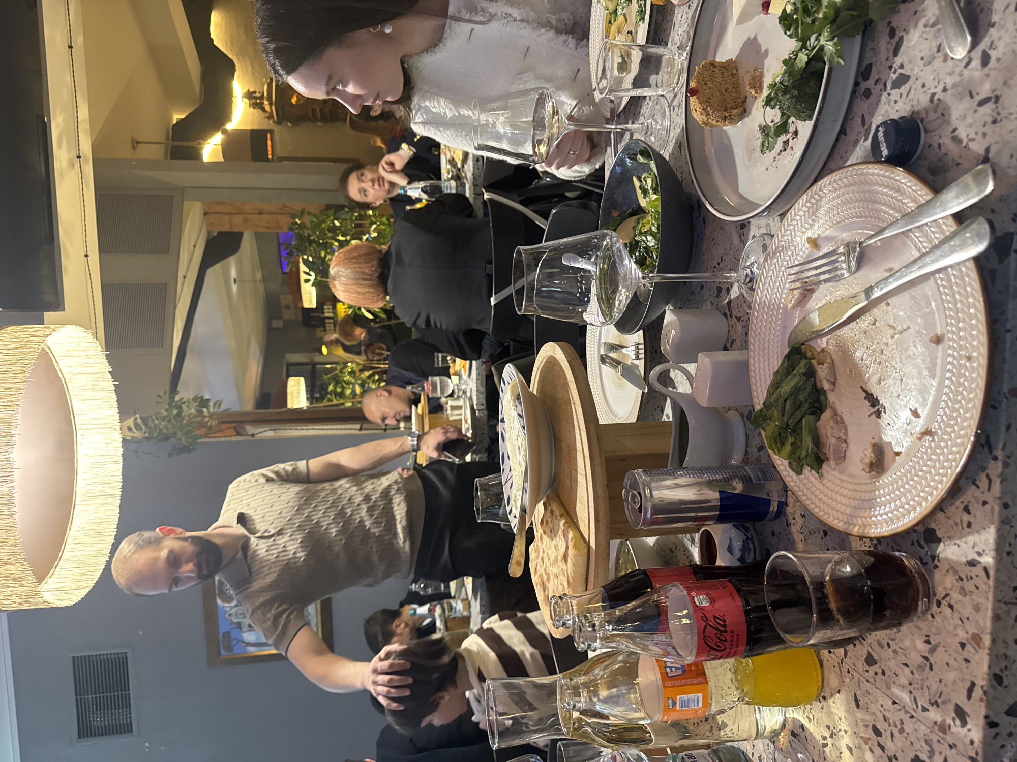 The image depicts a restaurant scene, likely in Tbilisi, Georgia. Diners are seated at a table laden with food and beverages, including finished plates, salad, bread, Coca-Cola, and Fanta. One man in a patterned beige shirt appears to be interacting with or touching the head of a seated person. Other individuals are visible at the table and in the background, engaged in conversation or dining. The restaurant has a modern interior with visible lighting fixtures and decorative elements.