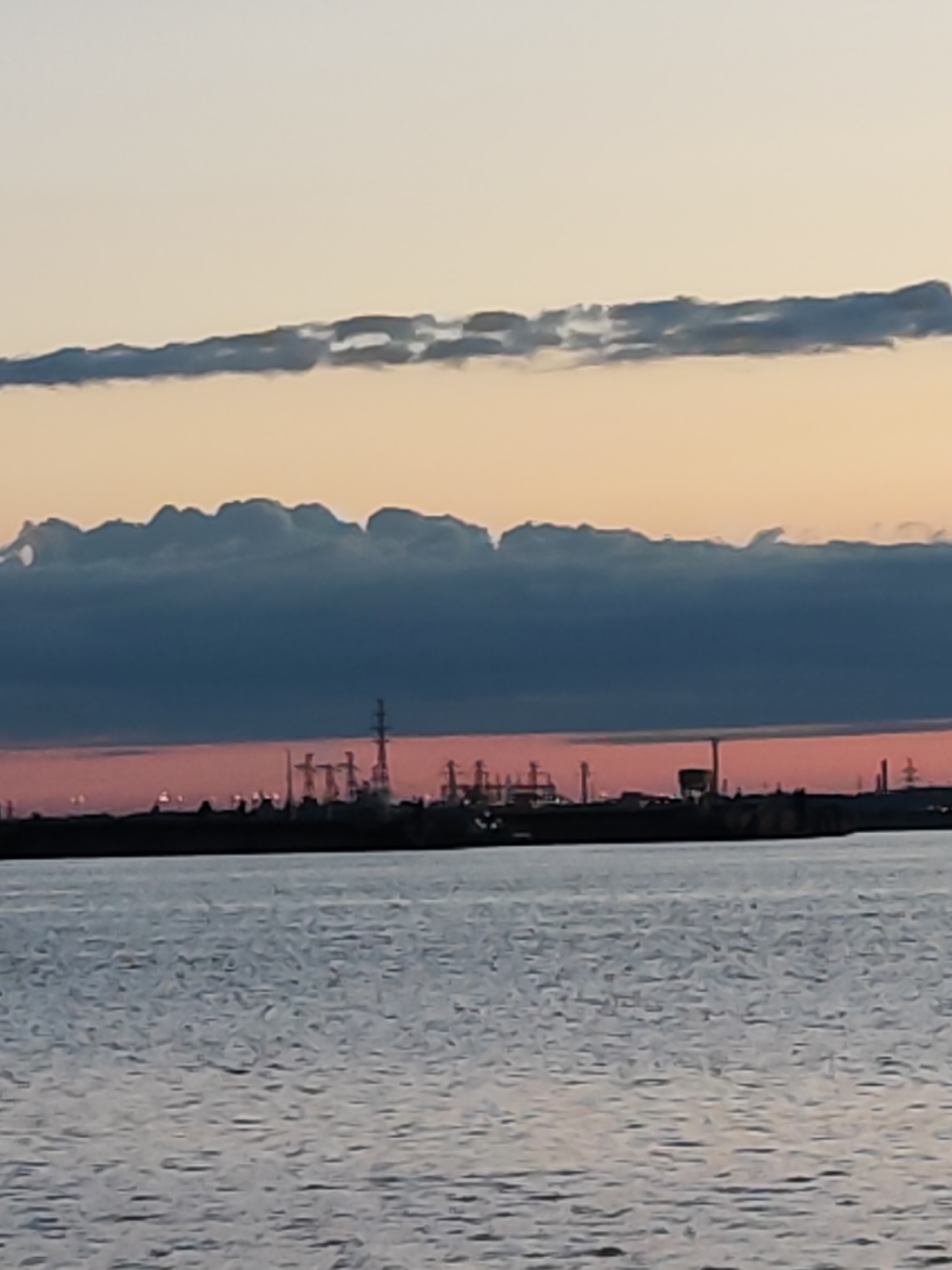 The image captures a twilight scene over a large body of water, likely a river or reservoir, with an industrial skyline in the distance. In the foreground, the water appears calm with subtle ripples, reflecting the ambient light. Across the midground, a dark silhouette of an industrial area stretches along the horizon, featuring numerous power line towers, factory buildings, and other industrial structures. Faint lights from the complex are visible against the sky.
The sky above presents a striking array of colors and cloud formations. A vibrant band of pink and orange hues glows just above the industrial skyline, indicative of either sunrise or sunset. Above this, a thick, dark grey cloud layer dominates the middle portion of the sky, its lower edge creating an undulating line. Higher up, a thinner, darker cloud band is visible against a pale orange background. The scene suggests a tranquil setting with a strong industrial presence, possibly in the vicinity of Chaikovsky, Russia. No people or visible text are present in the image.