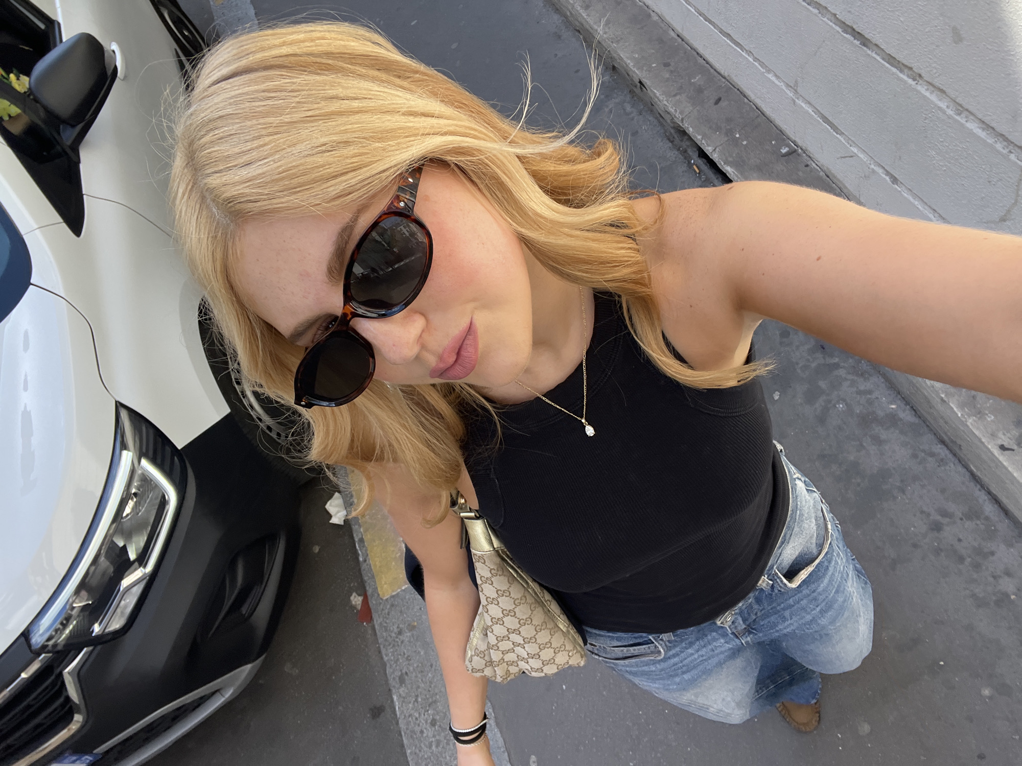 A woman is taking a selfie outside on a street in Paris, France. She is wearing sunglasses, a black tank top, blue jeans, and brown shoes. She has a gold necklace and a patterned purse on her shoulder. A white car is visible on the left side of the image. The street is gray and has a curb.
