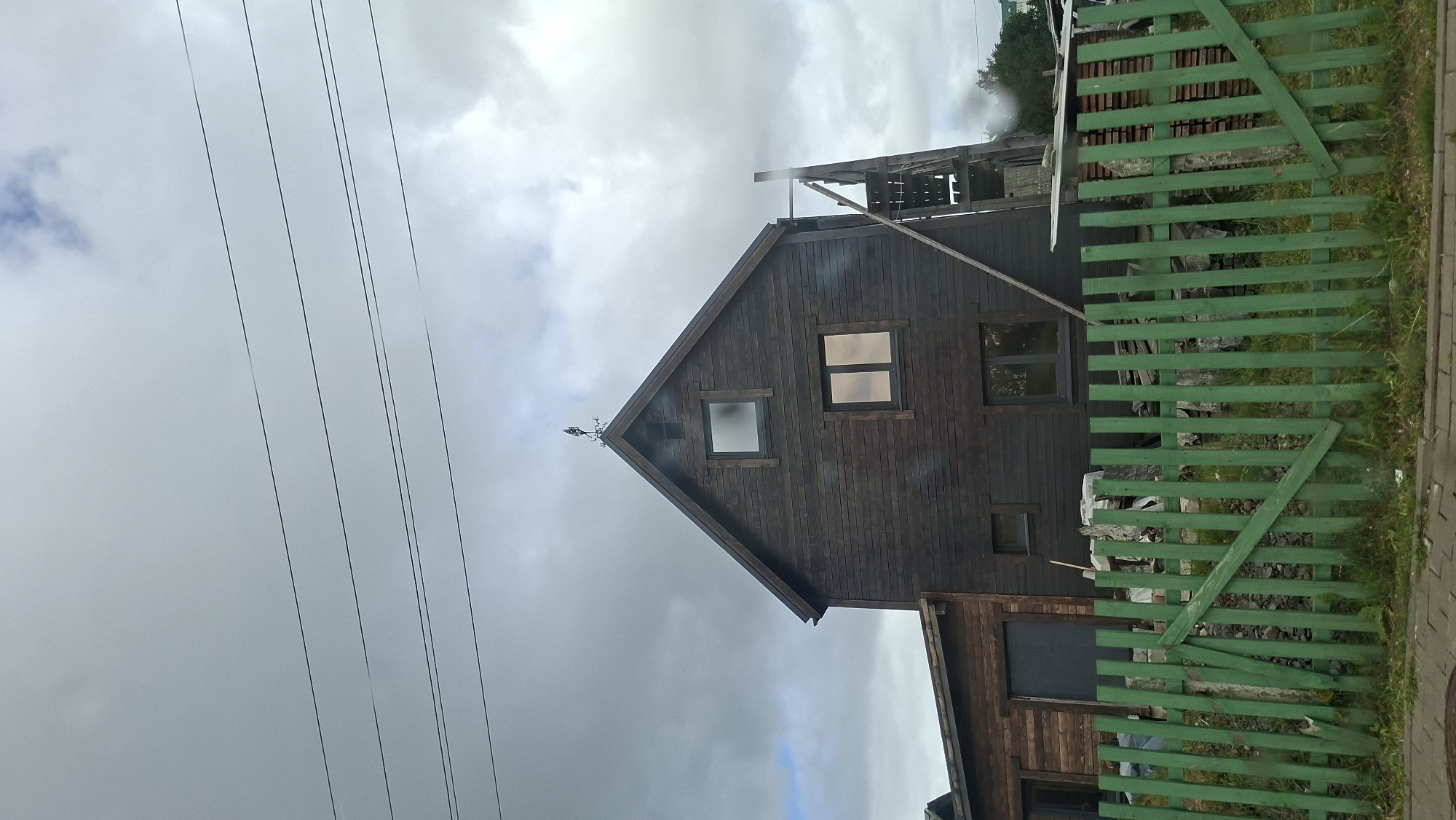 The image depicts a dark wooden house with a pitched roof and several windows. A weather vane, shaped like a bird, is visible at the apex of the roof. To the right of the house, a wooden staircase is leaning against the structure, suggesting ongoing construction or renovation. A bright green wooden fence runs across the foreground, with some wooden planks stacked behind it. A variety of overhead wires are also visible against the overcast sky. The scene appears to be in a rural or suburban area of Myadzyel, Belarus.