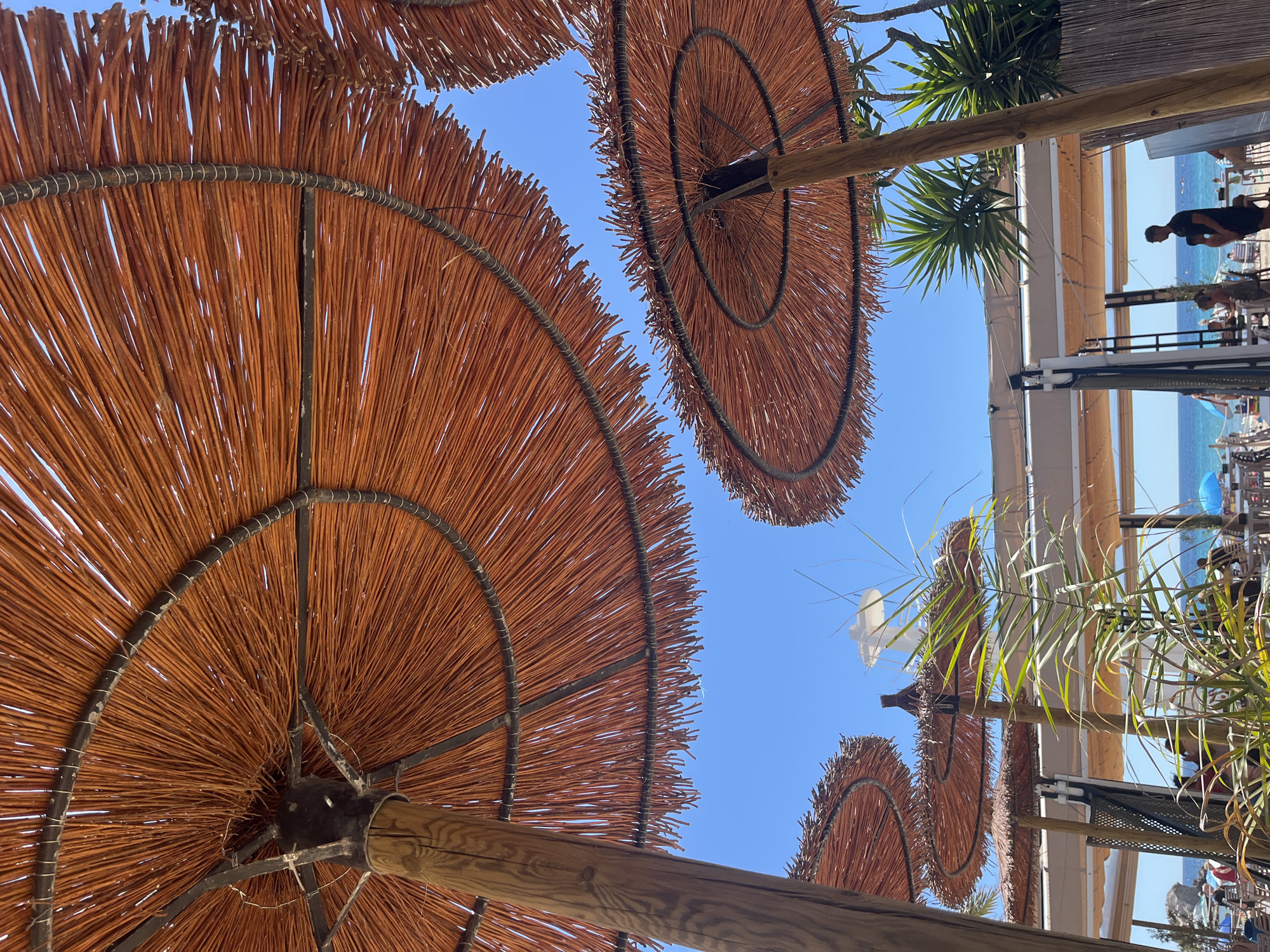 The image depicts a sunny scene in Teulada, Spain, looking upwards at straw beach umbrellas against a clear blue sky. The umbrellas are circular and made of dried straw, with visible wooden supports and metal rims. In the background, there is a glimpse of a beachside structure with people and palm trees. The ocean is also faintly visible in the distance.