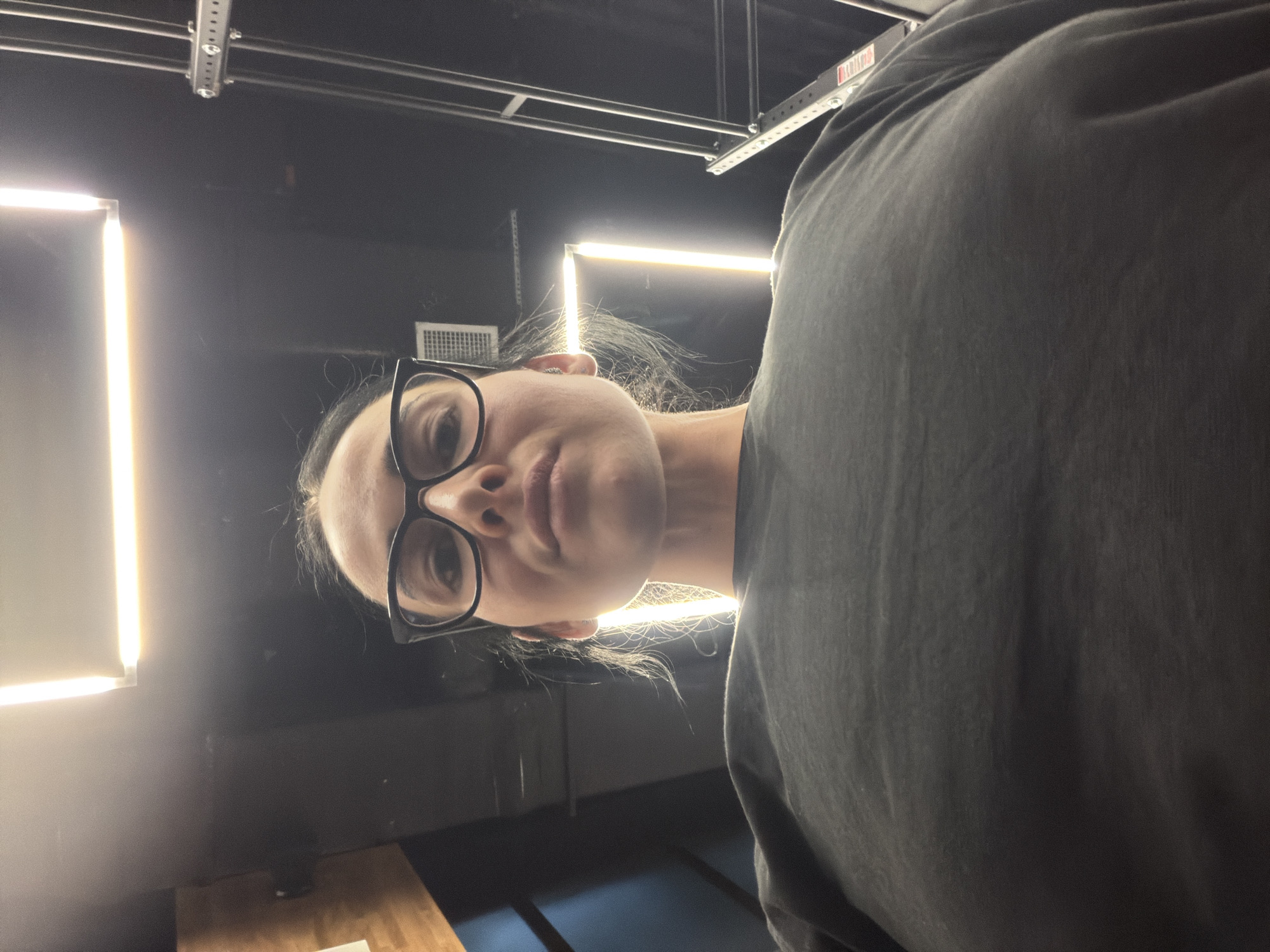 A low-angle shot features a person with pale skin and glasses, their face angled towards the camera. The person is wearing a dark grey, possibly oversized, garment that occupies a significant portion of the right side of the frame. The background reveals a dark, industrial-looking space with fluorescent tube lighting fixtures. The location is not discernible as being in Saint Petersburg, Russia, based on the visual information provided.