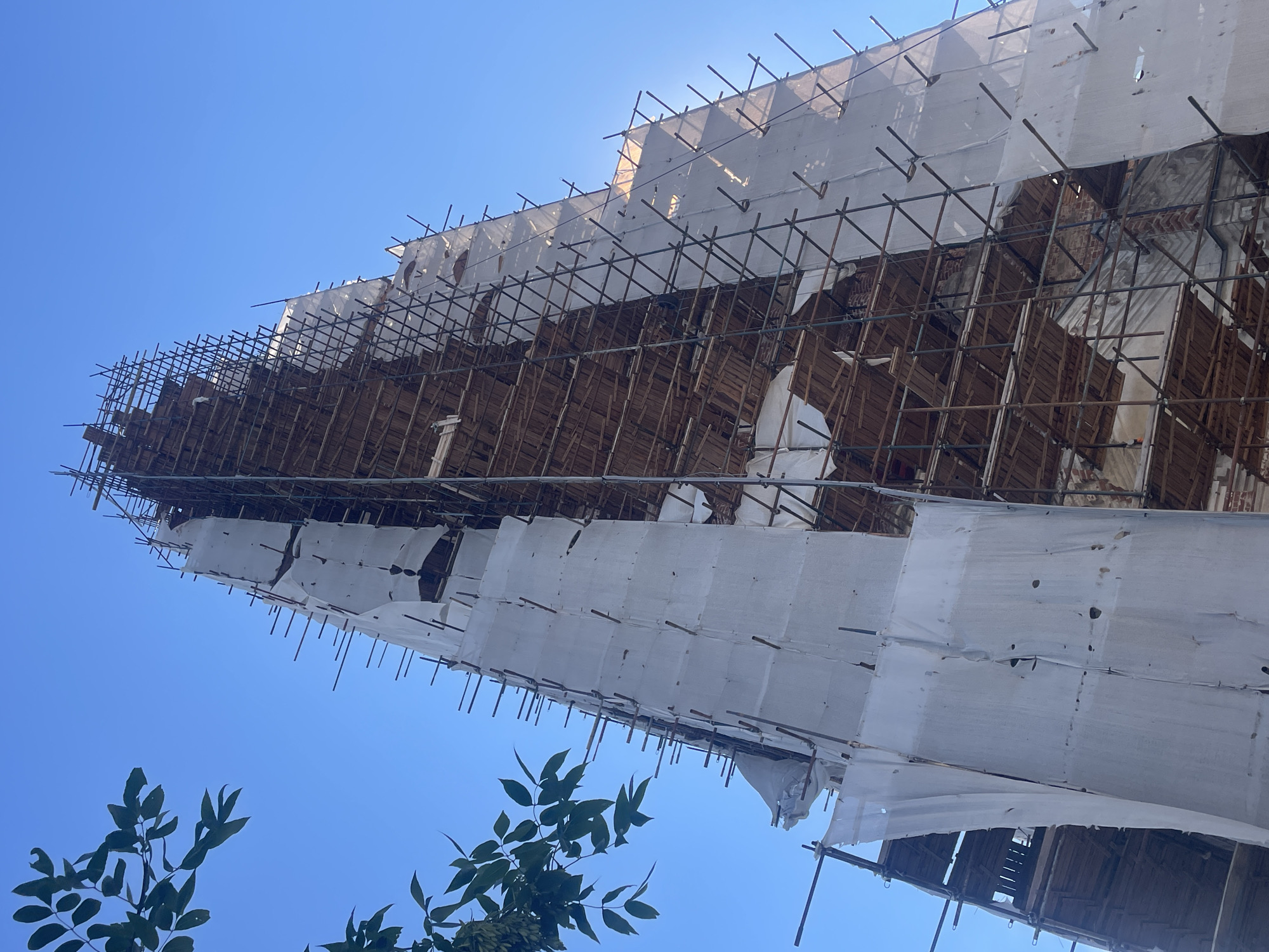 The image shows a low-angle view of a tall, spire-like structure, likely a church or bell tower, under renovation. The structure is covered in scaffolding made of metal poles and wooden planks. White tarps, some torn and weathered, are draped over the scaffolding, partially obscuring the underlying brickwork and wooden beams. The sky is a clear, bright blue. In the upper left corner, a branch with green leaves extends into the frame. This structure is located in Yuryevets, Russia.