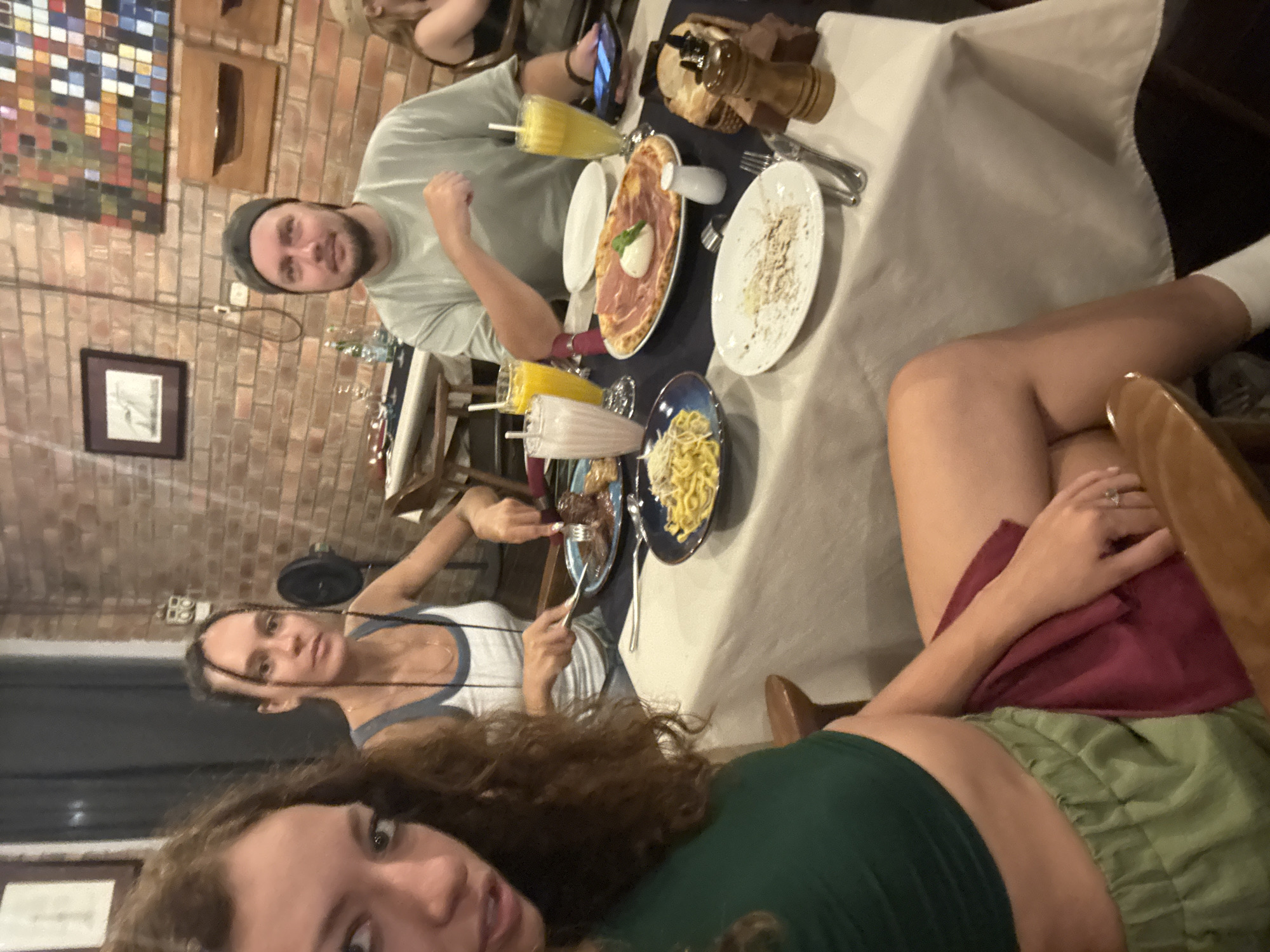 A group of four people are sitting at a table in what appears to be a restaurant. The table is covered with a white tablecloth, and several plates of food are visible, including a pizza with ham, a plate of pasta, and another plate with a white sauce. There are also glasses of juice on the table, as well as salt and pepper shakers. The people at the table appear to be enjoying themselves and interacting with one another. The background of the image shows a brick wall with a painting or photograph hanging on it.
Based on the style of the food and the overall atmosphere, it's possible that the restaurant is located in the city of Hà Nội, Vietnam.