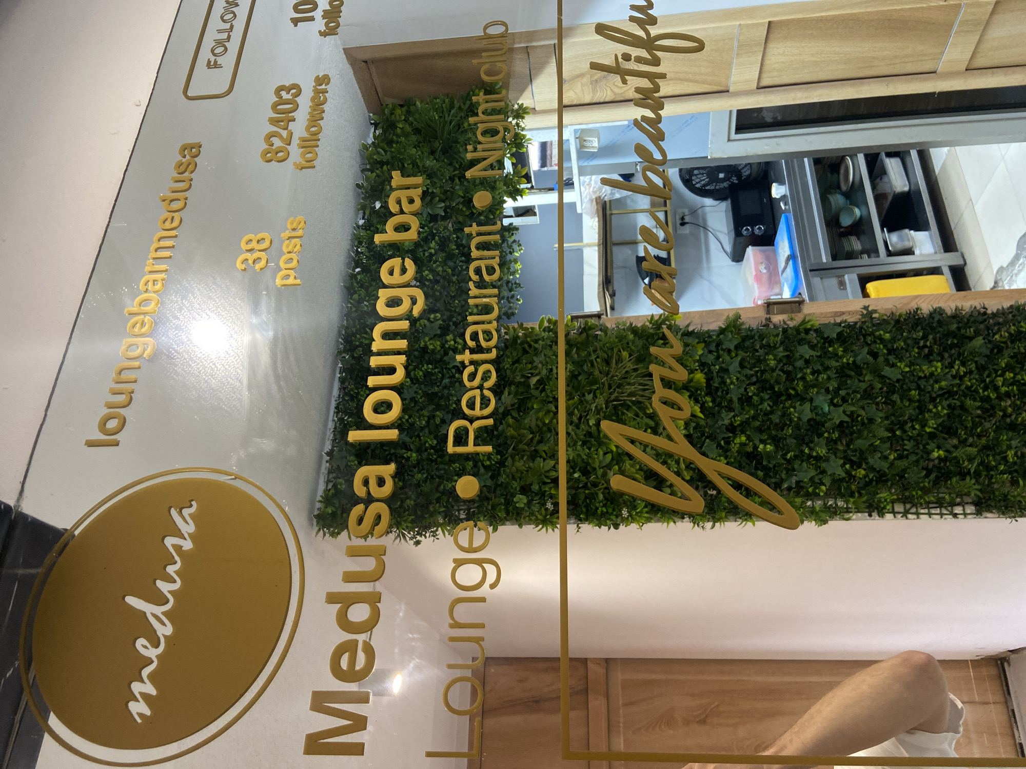 The image captures an interior view of a commercial establishment, likely an entrance or a decorated wall, located in Petrovac centar, Montenegro. The focal point is a clear, reflective surface, possibly glass, featuring golden 3D lettering and a logo.
The prominent text identifies the venue as "Medusa lounge bar," which also operates as a "Lounge," "Restaurant," and "Night club." A social media-inspired display is visible, showing "loungebarmedusa" (likely a handle) with a "FOLLOW" button, "38 posts," "82403 followers," and "10 follo" (presumably 'following'). The "Medusa" name is also presented within a distinct oval logo. Additionally, a cursive message reads "You are beautiful."
Behind the reflective surface, a vibrant artificial green plant wall creates a decorative backdrop. Beyond this wall, an internal space is visible, suggesting a functional area like a kitchen or service counter. This area includes metallic surfaces, drawers, various containers, and a dark appliance, possibly a coffee machine. Wooden paneling is also integrated into the background design. Part of a person's arm and leg, wearing light-colored clothing, is visible at the bottom right of the frame, either reflected or partially within the scene. The setting appears modern and inviting.