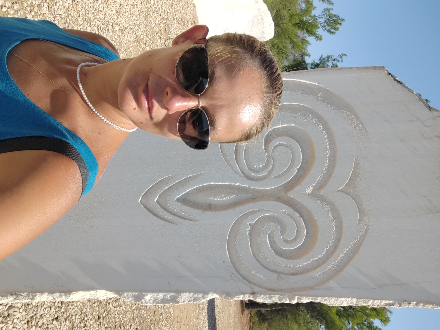 In Limenaria, Greece, a woman with blonde hair pulled back and wearing sunglasses, a blue tank top, and a pearl necklace poses for a selfie in front of a white stone sculpture featuring a swirling, abstract design. The sculpture is tall and roughly carved. The background consists of a bright blue sky, green trees, and a ground covered in small pebbles and gravel. The woman's face is turned towards the camera, and she appears to be smiling slightly.