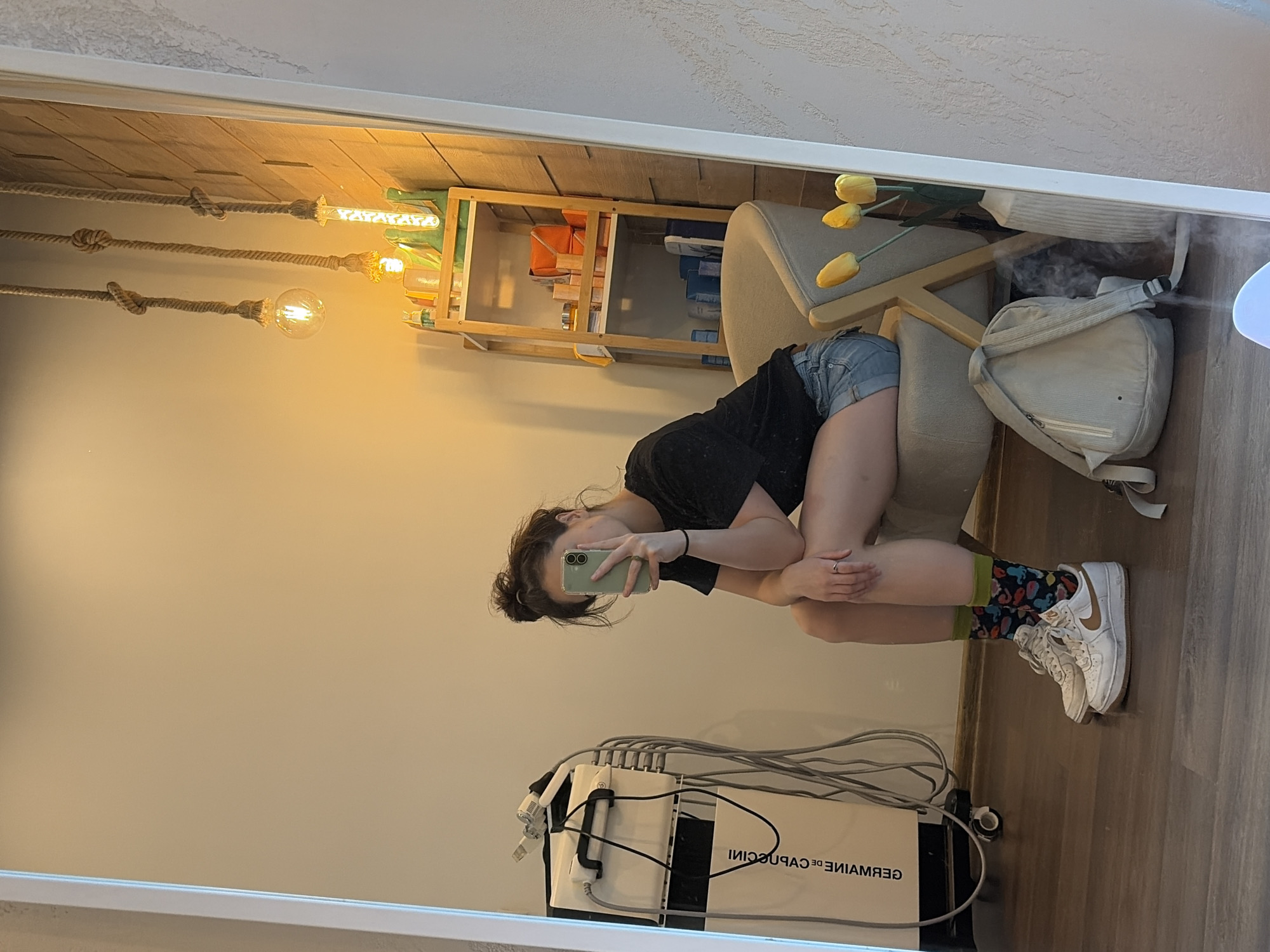 A person is captured in a mirror selfie, showcasing their lower body and midriff. They are seated on a chair, wearing denim shorts, a black t-shirt, and colorful, patterned socks with white sneakers. The person is holding a light green smartphone to take the photo.
The background reflects a room with wooden paneling on one wall, adorned with three pendant lights hanging from knotted ropes. To the right, a wooden shelf unit is visible, holding various items, including what appear to be orange containers and blue objects. A stack of yellow artificial tulips sits on a stool near the shelf.
On the floor, a piece of equipment with multiple tubes and cables is partially visible, bearing the text "GERMAINE DE CAPUCCINI" in black lettering. The overall lighting suggests it could be daytime or evening, with warm, artificial light illuminating the scene. The atmosphere appears casual and relaxed.