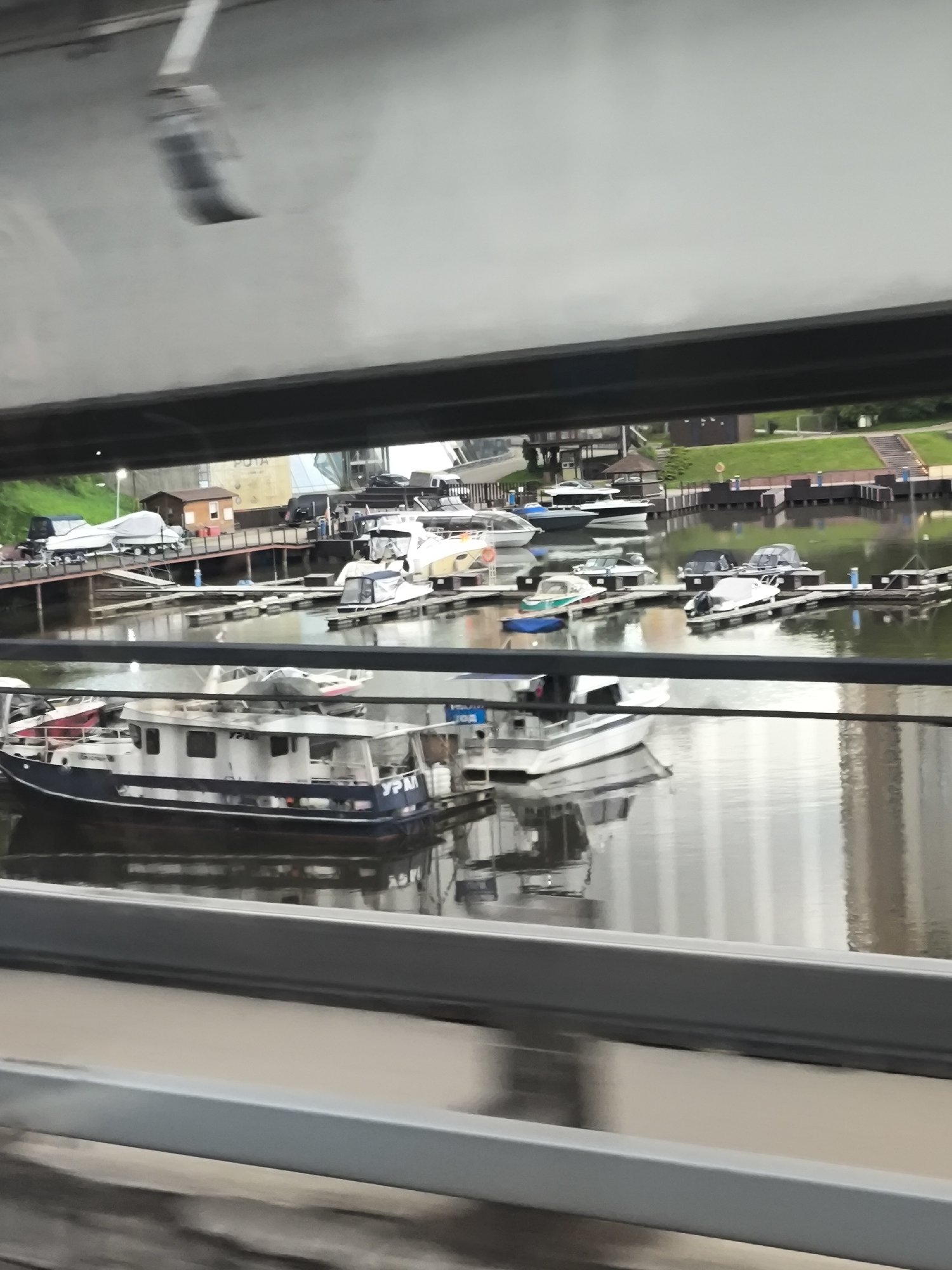 The image, taken from within a vehicle moving across a bridge or structure, captures a marina or harbor scene. The location is likely an urban area, as suggested by the presence of buildings reflected in the water. The foreground shows the interior structure of the vehicle from which the image was taken. Beyond this, there is a body of water with several boats docked along piers. The boats vary in size and type, including smaller recreational boats and a larger vessel with visible lettering "YPM" on its side. Across the water, a grassy bank is visible, rising to a higher elevation, with some buildings and structures nestled amongst the greenery.