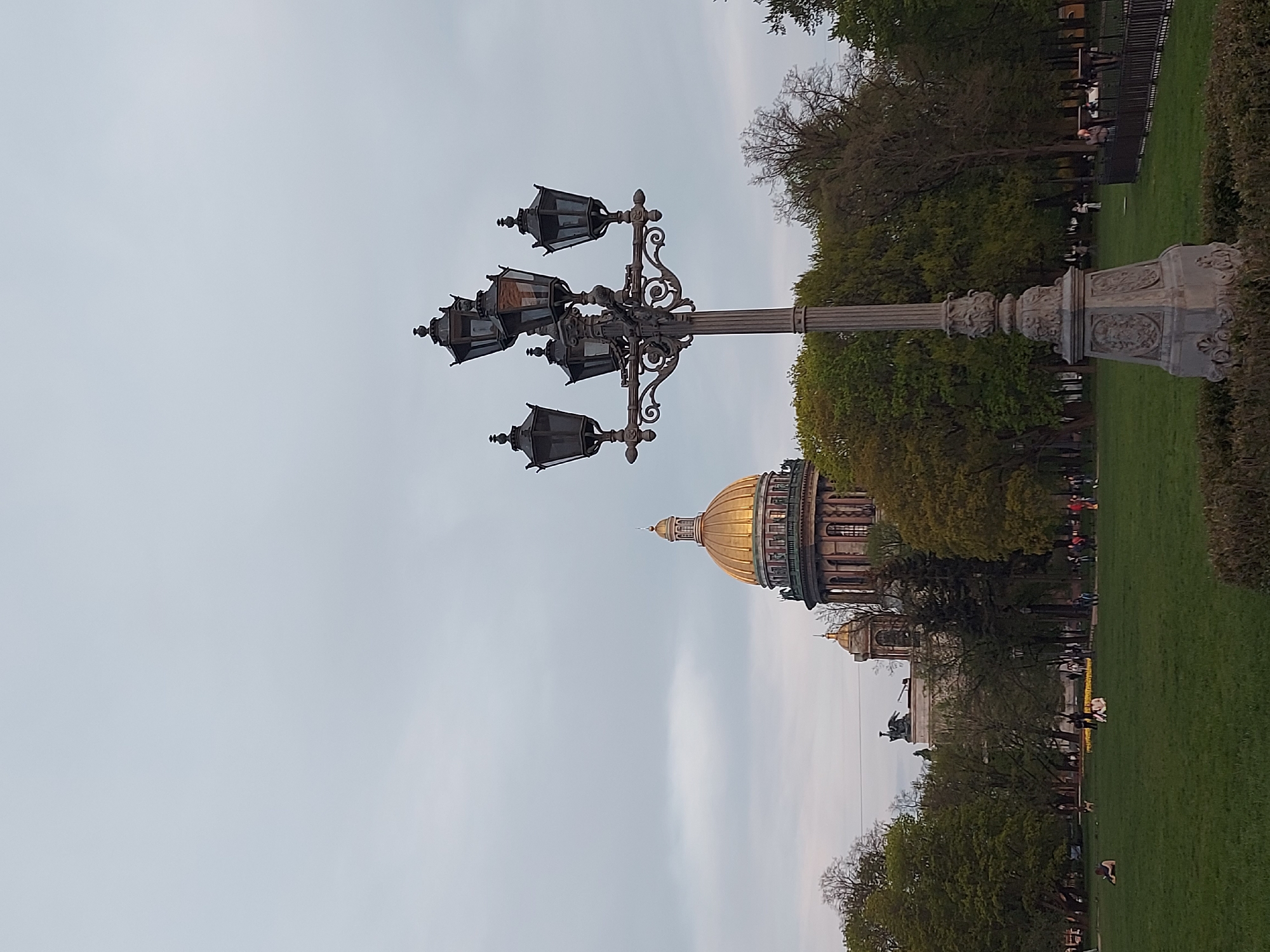 This image captures a scene in Saint Petersburg, Russia. The main focal point is a tall, ornate street lamp with multiple lanterns, standing in front of Saint Isaac's Cathedral, recognizable by its large gold dome. The foreground features green lawns and manicured hedges with people walking around. The sky is overcast, contributing to a muted lighting.