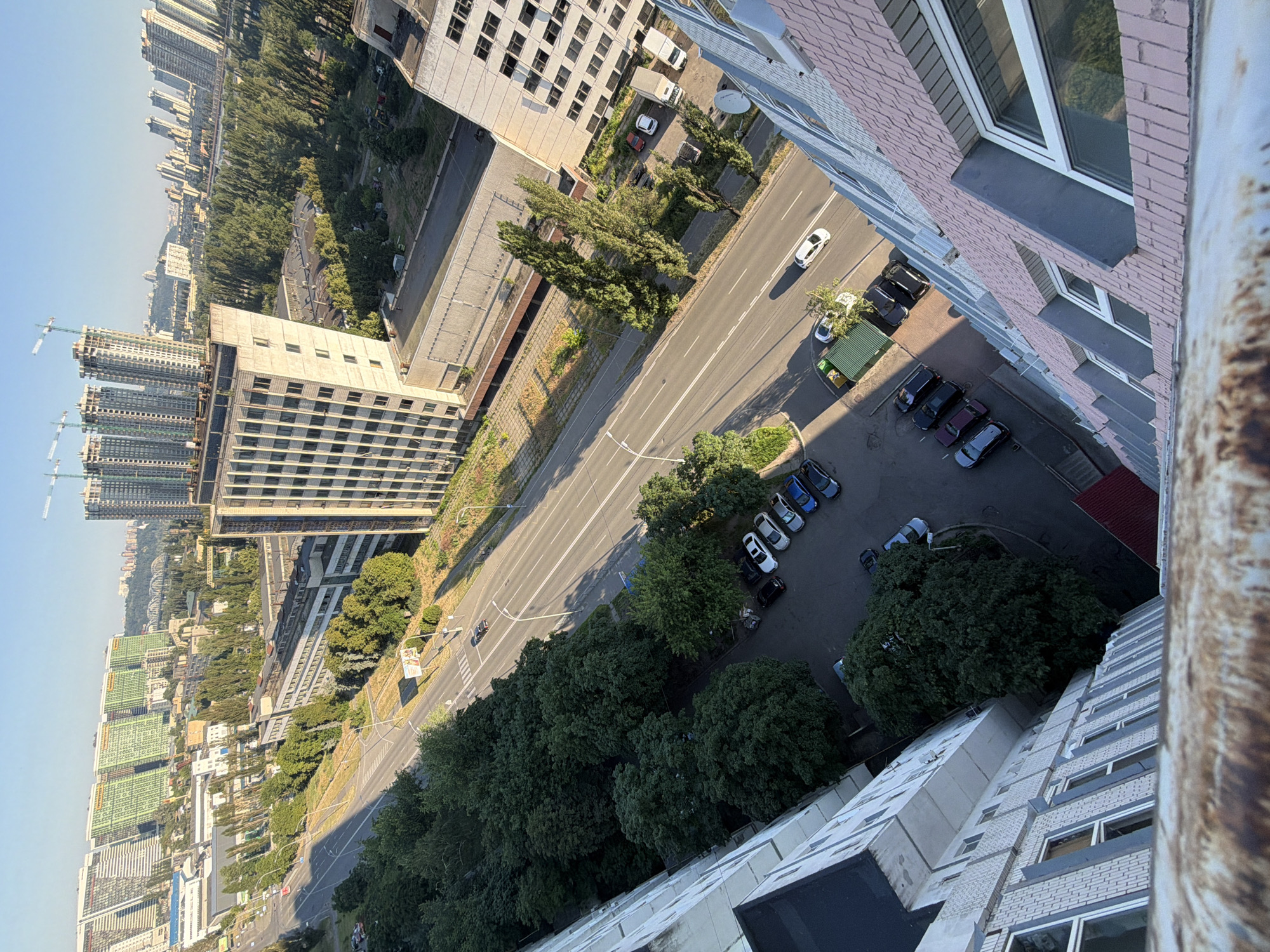 This is a high-angle, full shot image taken from a residential building overlooking a street and surrounding urban landscape in Kyiv, Ukraine. The foreground is dominated by the rust-colored, weathered metal railing of a balcony or rooftop. Below, the camera looks down on a multi-lane road with several cars parked on the side. To the left of the road, a dense line of mature green trees creates a natural barrier. Further beyond the trees, several large, uniform apartment buildings and industrial-style structures are visible. The background reveals a sprawling cityscape with more buildings, including modern high-rises under construction with cranes, and a bridge spanning a distant waterway. The sky is clear and blue, suggesting a sunny day. The scene depicts an everyday urban environment with a mix of residential and industrial elements.