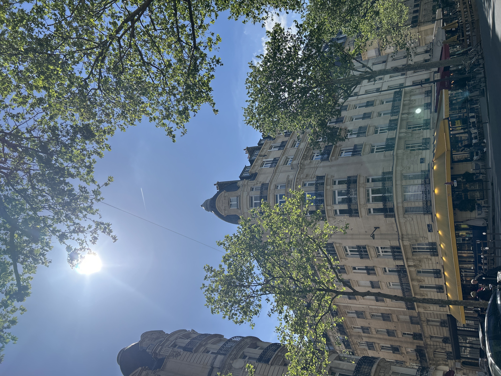 This media file is a low-angle photograph capturing a bright, sunny city street scene, primarily looking upwards towards the sky framed by buildings and trees.
The central subject is a clear, bright blue sky with a few wisps of white clouds. The sun is prominently featured in the lower left quadrant (when oriented correctly), casting a strong lens flare and illuminating the scene. A faint white contrail from an aircraft is visible in the sky.
Large, mature trees with abundant green leaves frame the upper and left edges of the sky, indicating a lush, possibly springtime or summer environment.
To the right and bottom of the frame, several multi-story buildings are visible. Their architecture is classical and ornate, characterized by light-colored stone facades, numerous windows with dark wrought-iron railings on balconies, and decorative elements. One building in the mid-ground features a distinctive rounded corner with a domed roof and attic windows, typical of Parisian Haussmannian design. Further down the street, other similar buildings line the thoroughfare.
At street level, parts of commercial establishments are visible, including a bright yellow awning and a red awning further down. Below the red awning, faint text "LE PONT..." can be partially discerned on a sign. Minimal street-level activity is hinted at by the presence of what appear to be restaurant or cafe tables and chairs under the awnings, and the tops of parked cars are visible at the very bottom edge of the frame.
The image suggests a pleasant, clear daytime setting in an urban European city, likely Paris, given the architectural style.