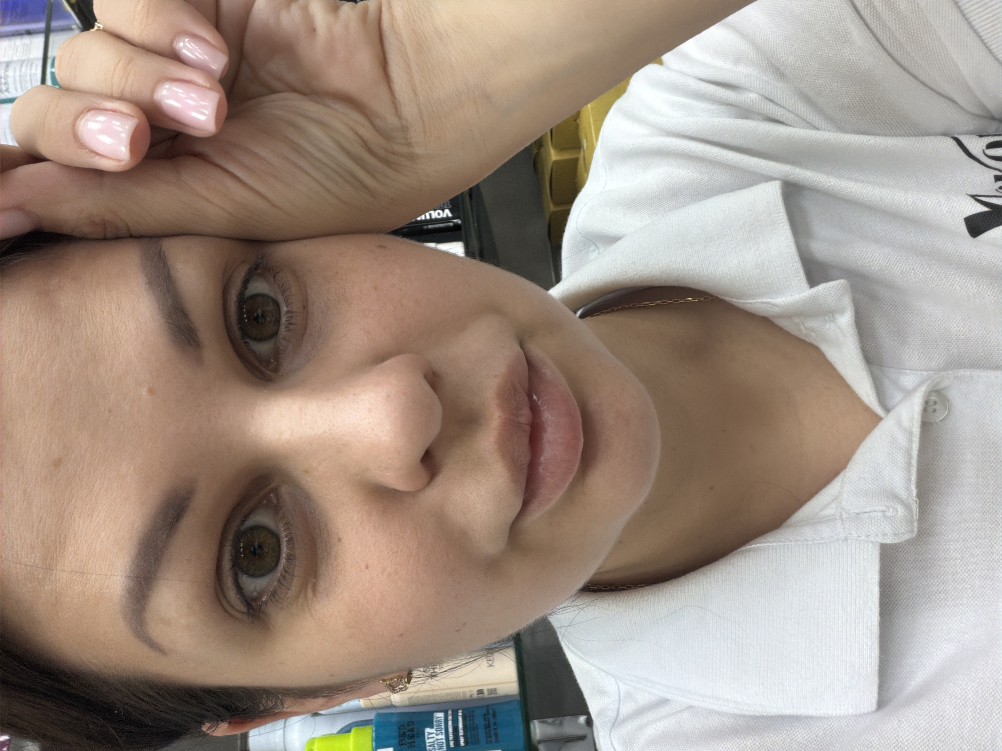 A close-up, portrait-oriented image shows a young woman indoors, with her head tilted and her left hand resting on her forehead. She has light skin, brown eyes, and is wearing a white collared shirt. A thin gold chain is visible around her neck. The background is blurred, but appears to include shelves with products, and the overall lighting suggests an indoor setting, possibly a store or salon. The woman's expression is neutral, and her fingernails are painted a pale pink. The orientation of the image is such that the woman's face is approximately horizontal, with her hair framing the bottom of the frame. Some text is visible on products in the background, including "RED HEAD" and "SALTY NOT SORRY". The context provided is Sochi, Russia.