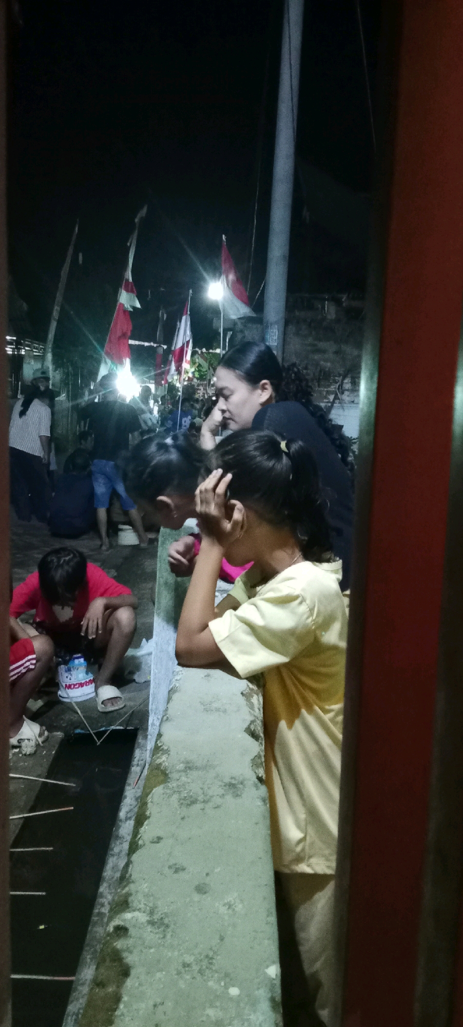 In an outdoor urban setting at night, likely in Kesamben, Indonesia, a group of people is gathered. In the foreground, two young girls are partially visible, one in a yellow shirt with her hand on her head, and another to her left also with her hand near her face. To the left of the girls, a young boy wearing a red shirt and red shorts is crouching, looking down at something on the ground. He is wearing white sandals. In the background, several adults are visible, some standing and some sitting. Red and white flags, possibly national flags, are displayed on poles, illuminated by artificial lights. A utility pole stands prominently to the right of the frame. The ground appears to be a rough concrete surface, and there is a dark, shallow channel of water running horizontally across the lower part of the image. The overall scene suggests a community gathering or event taking place after dark.