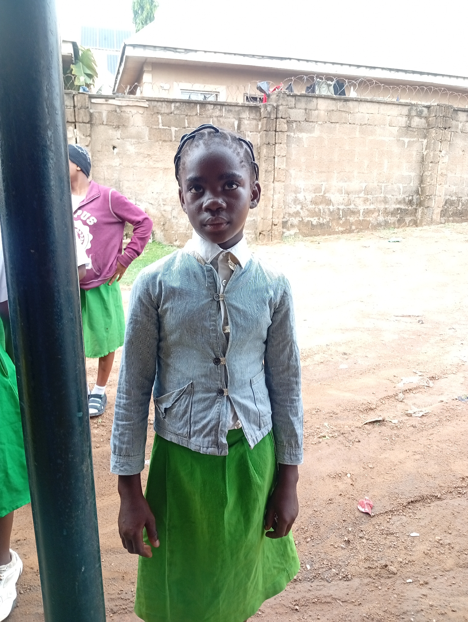 A young girl stands centered in the frame, dressed in a school uniform consisting of a green skirt and a light blue striped jacket over a white collared shirt. Her hair is braided and pulled back from her face. Behind her is a cinder block wall topped with barbed wire. To her left, partially obscured by a black pole, another girl is visible wearing a green skirt and a purple jacket. The ground appears to be dirt or mud. The location is Narayi, Nigeria, as the prompt requests.
