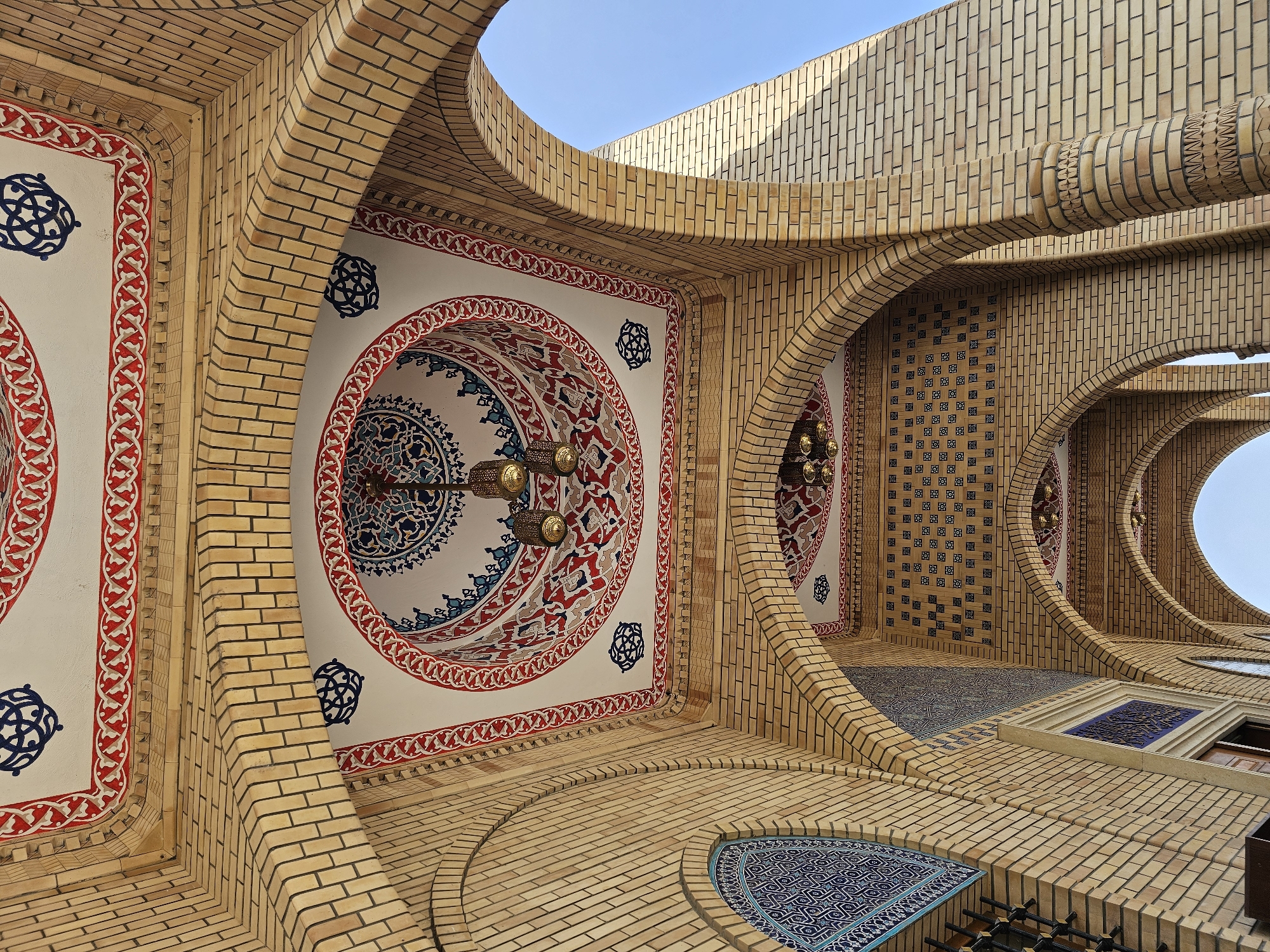 The image shows a low-angle view of the interior of an arched passageway with a decorative ceiling. The passageway is constructed from light-colored bricks. The ceiling is painted white and features intricate geometric patterns in red, blue, and black. Hanging from the ceiling are three ornate metal lanterns. In the background, a clear blue sky is visible. The style of the architecture and decoration suggests a location with Islamic cultural influence. While the query specifies Özgön, Kyrgyzstan, this visual information alone does not confirm the location.