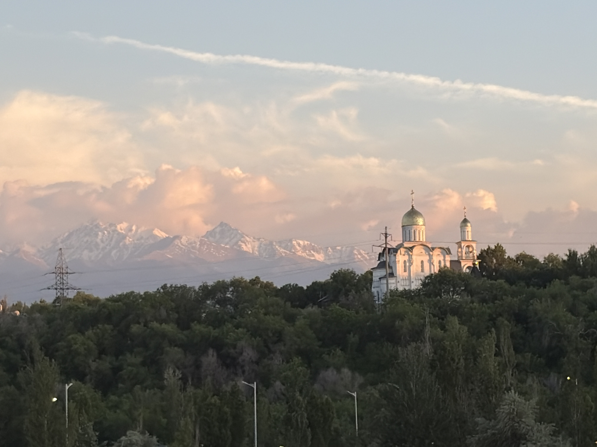 The image shows an Orthodox church in Almaty, Kazakhstan, situated on a hill covered in green trees. The church is white with golden domes, and stands out against the backdrop of the hill. Behind the hill are snow-capped mountains, partially obscured by light pink clouds. In the foreground, there are trees, streetlights, and a power line pole. The sky is a mix of light blue and pink, suggesting either sunrise or sunset.