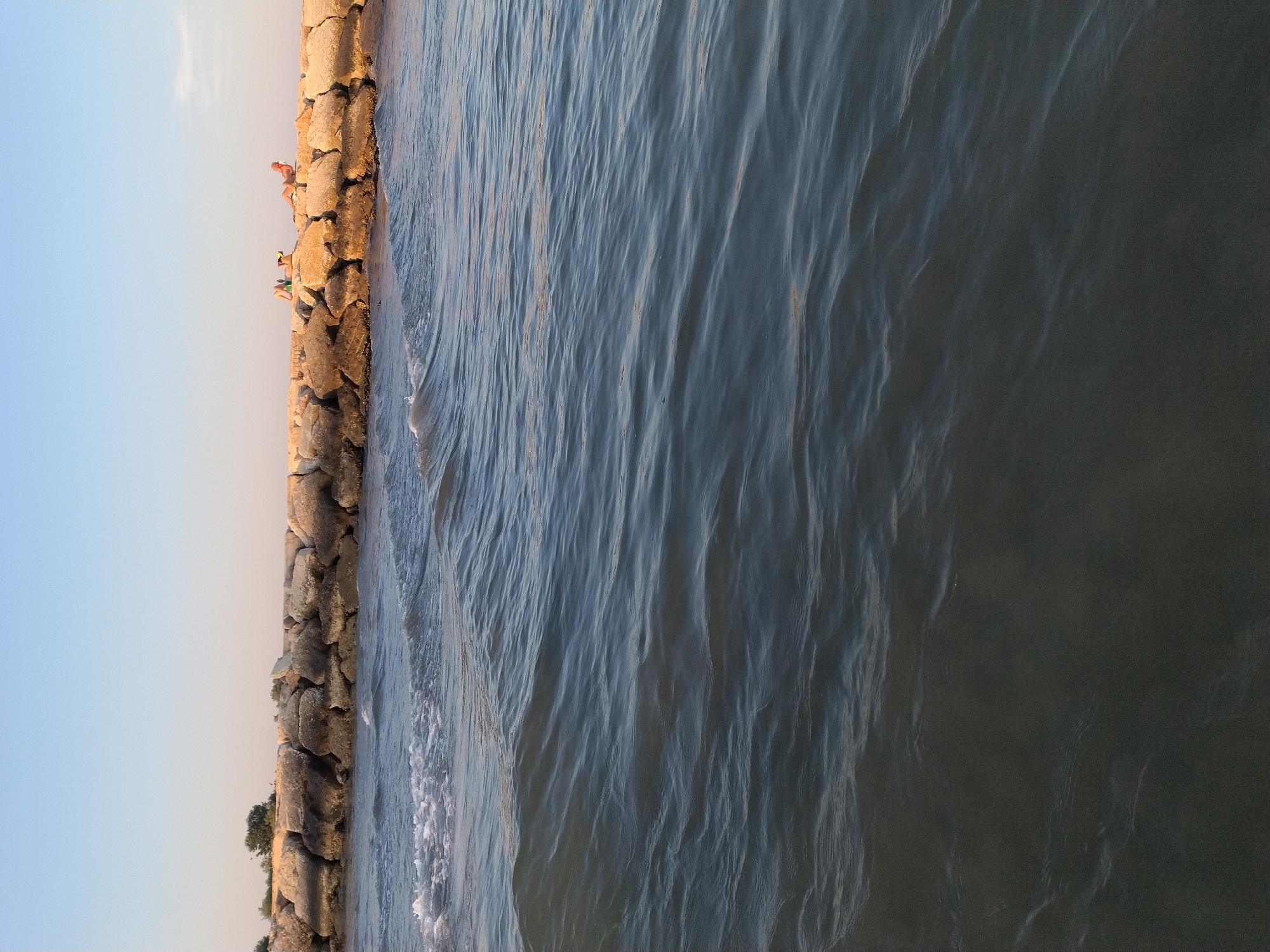The image shows a breakwater made of large stones, with two people sunbathing on top. The breakwater is in the sea, and there are waves in the foreground. The sky is clear and blue. The breakwater is located in Caorle, Italy. The two people are on the breakwater. One person is lying on their back, and the other is sitting up. They are both wearing swimwear. The sea is calm, with small waves. The breakwater is made of large, rough stones. The sky is clear and blue, with a few wispy clouds. The overall impression is one of a peaceful summer day at the beach.