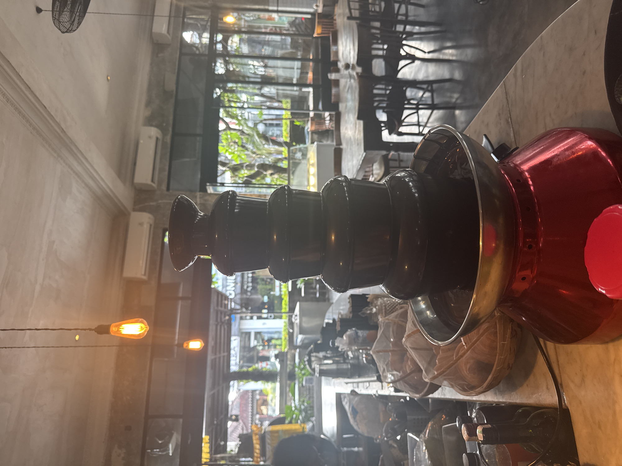 The image shows a chocolate fountain on a marble table in what appears to be a restaurant or cafe. The fountain is primarily black, with a stainless steel bowl at the top and a red base. The background features a blurred view of the restaurant interior, including tables, chairs, large windows, and lighting fixtures. Based on the information available, the location is in North Kuta, Indonesia.