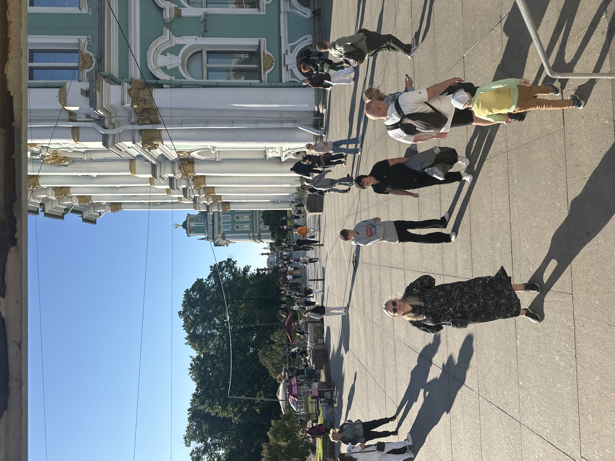 The image shows a wide, paved plaza in front of a large, ornate, pale teal and white building with golden decorative elements and tall columns. The building appears to be the Winter Palace on Palace Square in Saint Petersburg, Russia. Sunlight casts long shadows across the plaza, indicating a sunny day. A group of tourists are walking around the plaza. In the foreground, a woman in a black and white patterned dress stands facing the camera, with a black crossbody bag. To her left, two women in casual clothing are walking. Further back, a group of people are visible, some walking, some standing. There are also two food kiosks with umbrellas in the mid-ground. In the background, trees and other buildings, including a green and gold spire, are visible against a clear blue sky. The overall scene is one of a bustling tourist attraction on a bright, sunny day.