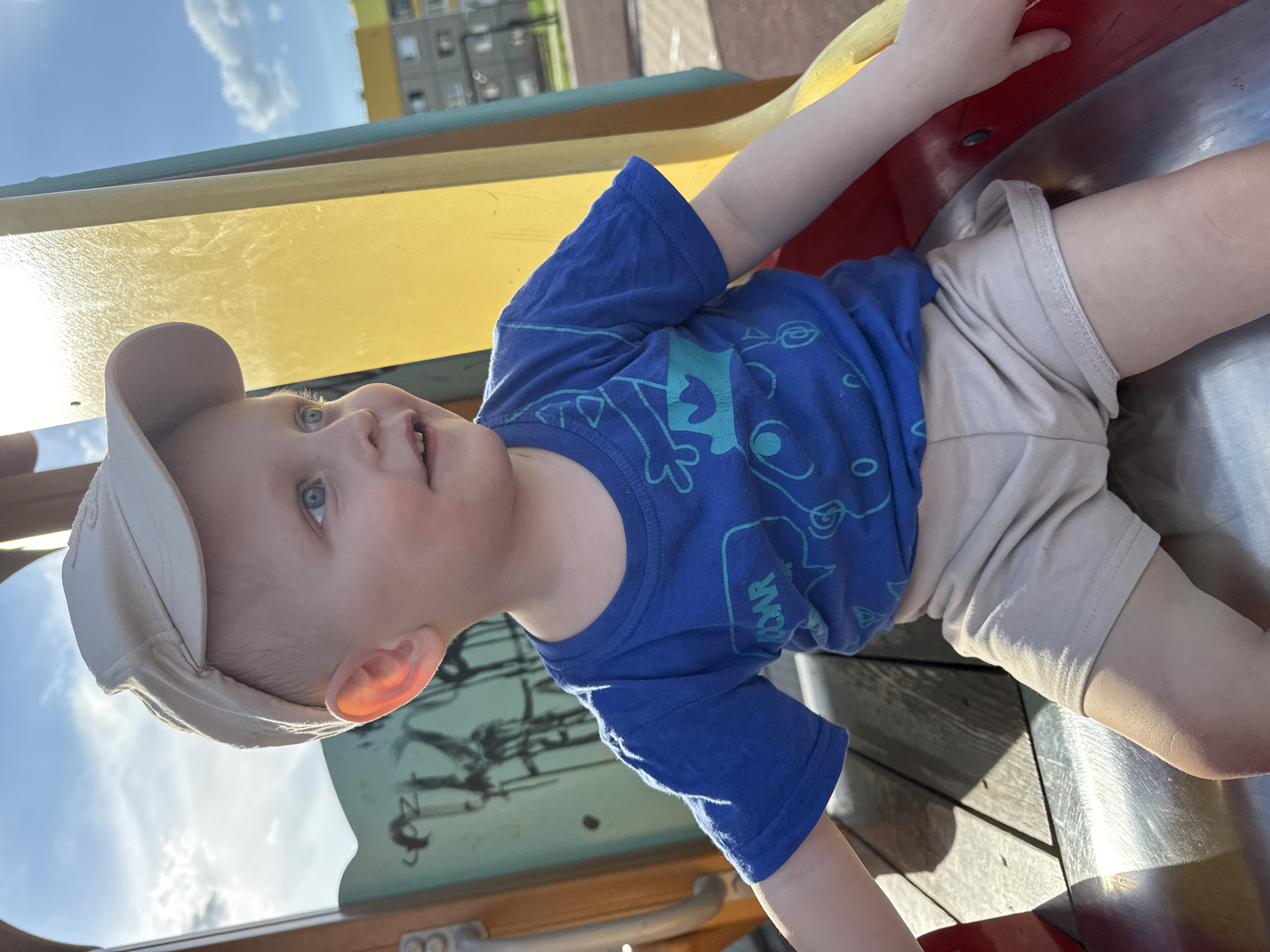 A young child with fair skin, wearing a light-colored baseball cap, a blue t-shirt with turquoise-colored graphic designs, and light-colored shorts, is lying on a playground slide. The slide appears to be made of metal and plastic, with visible red, yellow, and green sections. In the background, the sky is visible with a few clouds and a multi-story residential building with yellow and gray sections. The location is in the city of Марковский, Russia.