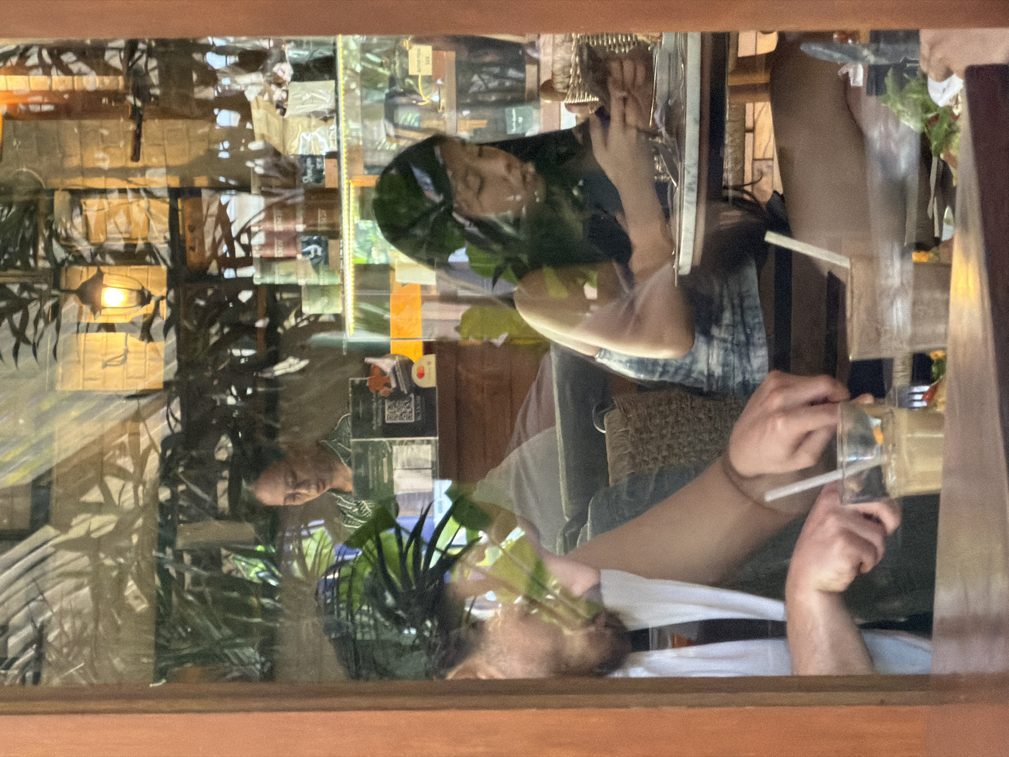 The image depicts a reflection on a window, likely at a cafe or restaurant in North Kuta, Indonesia, given the presence of tropical foliage and architectural elements consistent with the region. In the foreground, the reflection shows a person's hands, one holding a utensil and actively cutting into food on a plate, with a glass containing a beverage and a straw nearby. Partially visible is another person wearing a white tank top. In the middle ground, the reflection shows a woman with dark hair, wearing a dark top and denim shorts, looking down at a smartphone. Behind her, another woman with glasses, wearing a patterned shirt, is visible behind a counter, possibly serving. The background reveals shelves stocked with various items, including bottles and packaged goods, and the warm glow of a lamp. The scene suggests a casual dining or social gathering.