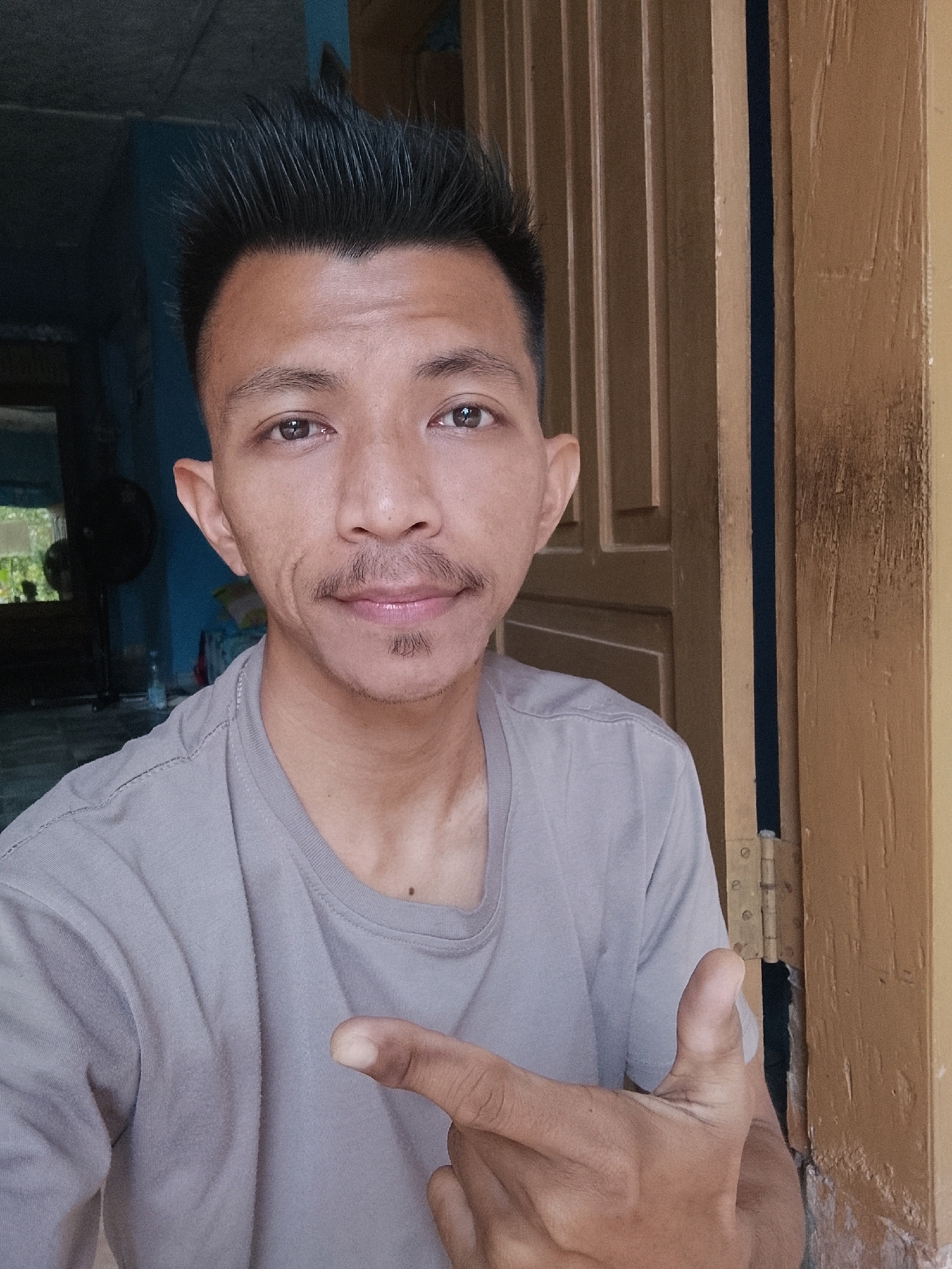 A medium shot depicts a young man of Southeast Asian descent in Kertahayu, Indonesia, posing for a selfie. He has short, spiky dark hair, a thin mustache, and a small goatee. His eyes are brown and his skin is tanned. He is wearing a light gray t-shirt and pointing his right index finger towards the left of the frame. The background shows an interior space with a blue wall on the left, a black standing fan, and a window with greenery visible. On the right, a light brown wooden door frame with visible hinges suggests the man is standing in a doorway. The lighting is natural and appears to be coming from the front.