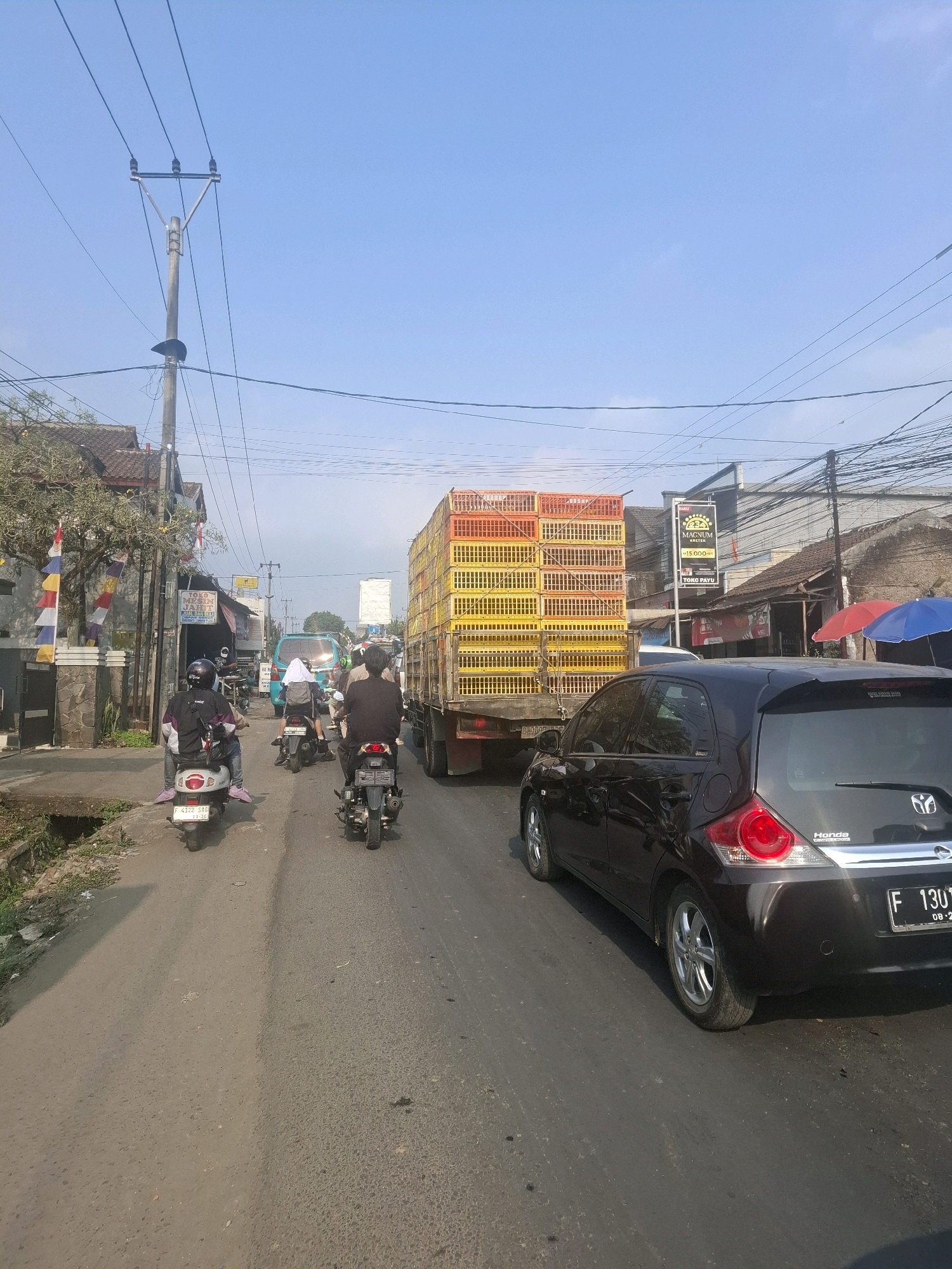 The image captures a street scene in Karangtengah, Indonesia, characterized by moderate traffic under a clear blue sky. A black Honda hatchback is prominently featured in the foreground, facing away from the viewer. To its left, a large truck is parked, its cargo area stacked with numerous yellow and orange plastic crates, likely used for transporting poultry. Several motorcycles are also present, with riders navigating the road. One rider on a white scooter is visible from behind, wearing a dark jacket and helmet. Another motorcycle, also seen from behind, is directly in front of the Honda. Further down the road, a turquoise van with a white cloth draped over its rear is observed.
The environment suggests a commercial or semi-urban area with buildings lining the street. Some buildings appear to be shops, indicated by signage. A power pole with electrical wires stretches across the upper portion of the image, a common sight in Indonesian streetscapes. The road surface is asphalt, showing signs of wear and tear, and is flanked by a drainage ditch on the left side. The overall impression is of a typical day with everyday traffic flow in a localized Indonesian town.