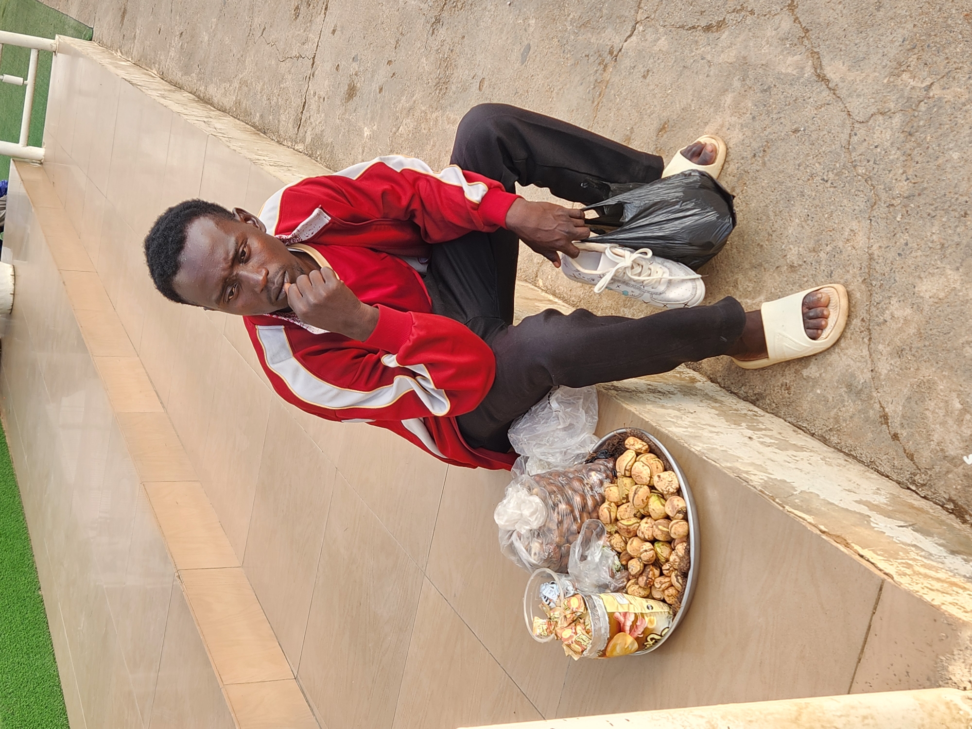 A person is sitting on a light-colored stone ledge, wearing black pants, a red jacket with white stripes, and tan sandals. They are looking down and touching their chin with one hand. Next to them is a black plastic bag, on top of which is a pair of white sneakers. In front of them is a round tray filled with assorted wrapped candies and what appear to be nuts in plastic bags. The setting appears to be outdoors, near a wall with green artificial turf on the other side of a white railing. The city and specific location are unknown.