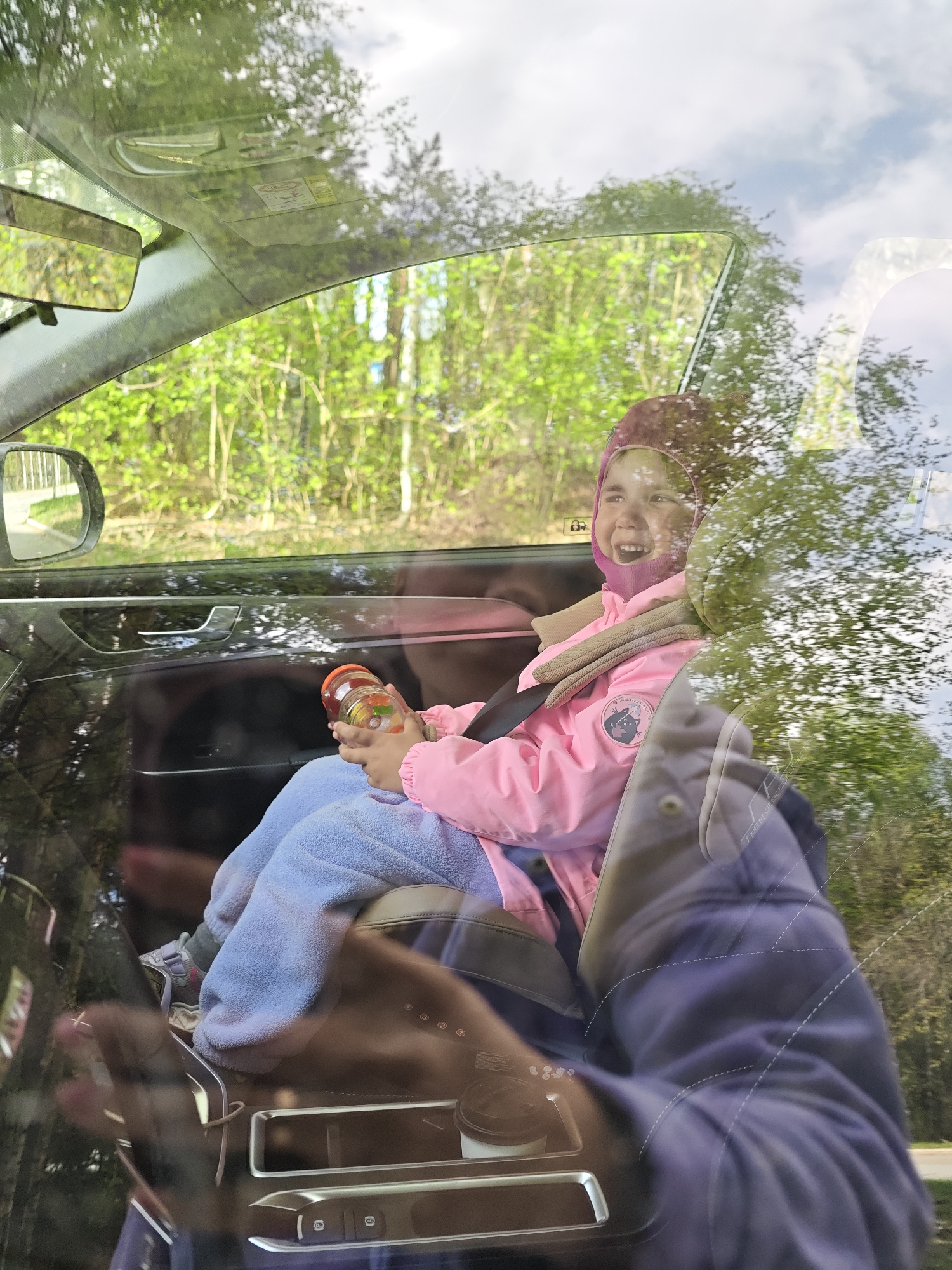 A young girl is seated in the back of a car, buckled in and holding a jar. She is wearing a pink jacket and a hat. The scene appears to be taken in a car moving near a wooded area, viewable through the car window. It is not possible to confirm the exact location of Chaikovsky, Russia, or any further specific location details from this image.