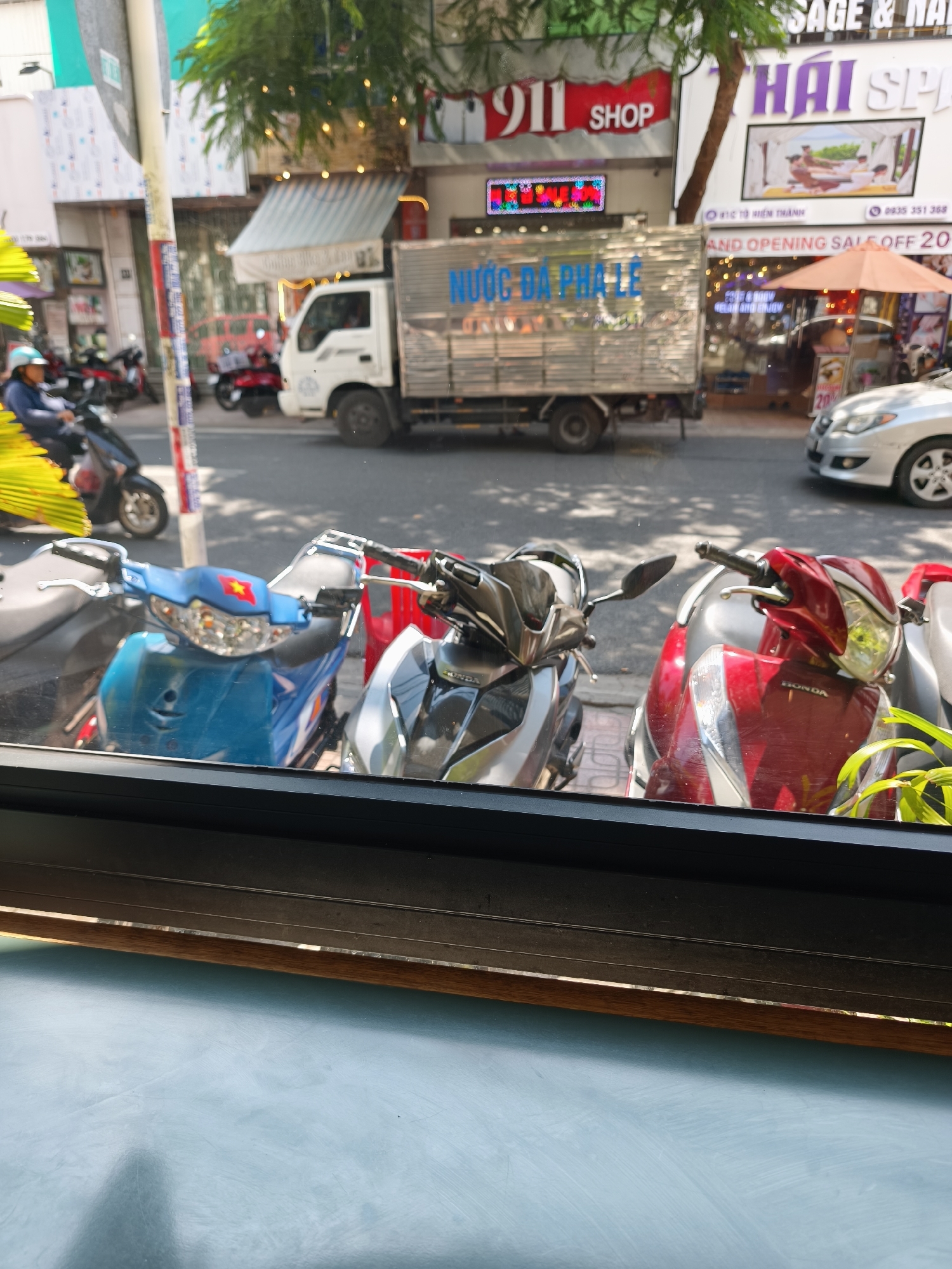 In Nha Trang Ward, Vietnam, a street scene unfolds with a variety of vehicles and businesses. In the foreground, three motorcycles are parked, one blue with a red star emblem on its front, one silver, and one red. Behind them, a white truck with "NƯỚC ĐÁ PHA LÊ" (iced tea) printed on its side is stopped. Further down the street, a silver car is parked. Businesses line the street, including one with "911 SHOP" signage and another that appears to be a spa, indicated by "THAI SPA" and an advertisement depicting people receiving massages. The environment suggests a sunny day, with shadows cast on the pavement. A person wearing a blue shirt and helmet is visible riding a motorcycle in the background.