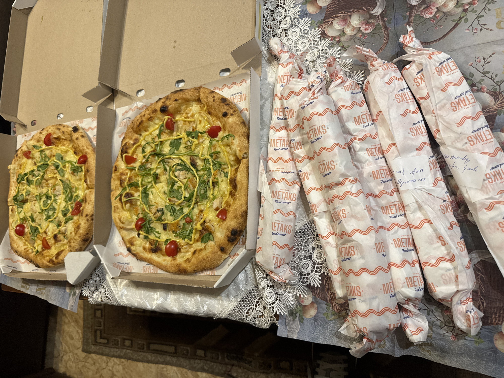 Here are two pizzas in open cardboard boxes and five food wraps from "METAKS Food Corner," which, according to its logo, was established in 2015. The location is likely in Yerevan, Armenia, due to the METAKS branding. The pizzas appear to have a thin crust and are topped with ingredients like tomatoes, greens, and a yellowish sauce. The food wraps are packaged in white paper with a red wavy pattern and the "METAKS Food Corner" logo printed repeatedly. They are lying on a patterned surface that might be a tablecloth. The scene is indoors, possibly on a dining table.