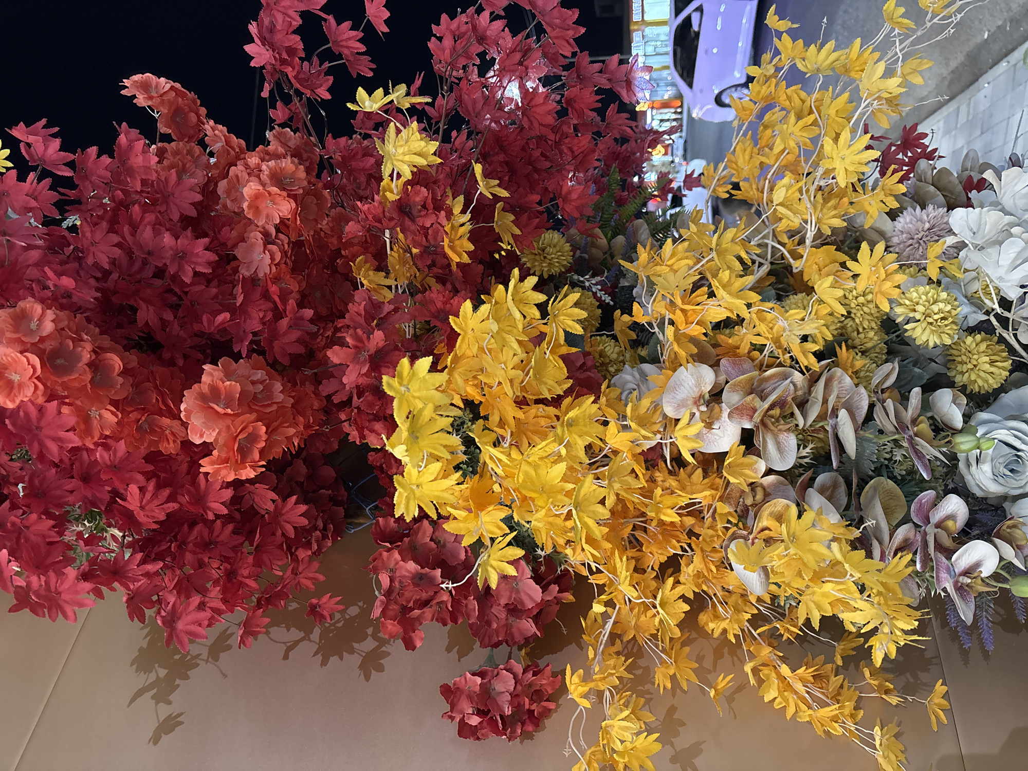 The image displays an outdoor floral arrangement in the city of Shymkent, Kazakhstan. The arrangement consists of artificial foliage and flowers in vibrant red and yellow hues, with some autumnal colors like orange and brown also present. The red foliage, resembling maple leaves, dominates the upper portion of the arrangement, while yellow maple-like leaves cascade downwards. Interspersed within the foliage are various artificial flowers, including orchids in shades of beige and purple, and larger blooms in muted tones of grey and white at the bottom. The arrangement is set against a neutral-colored wall on the left, casting shadows of leaves and flowers onto it. To the right, a dimly lit urban street scene is visible, with a light purple car parked and illuminated storefronts in the background. The lighting suggests it is nighttime. There are no people clearly visible in the immediate foreground or interacting with the floral arrangement.