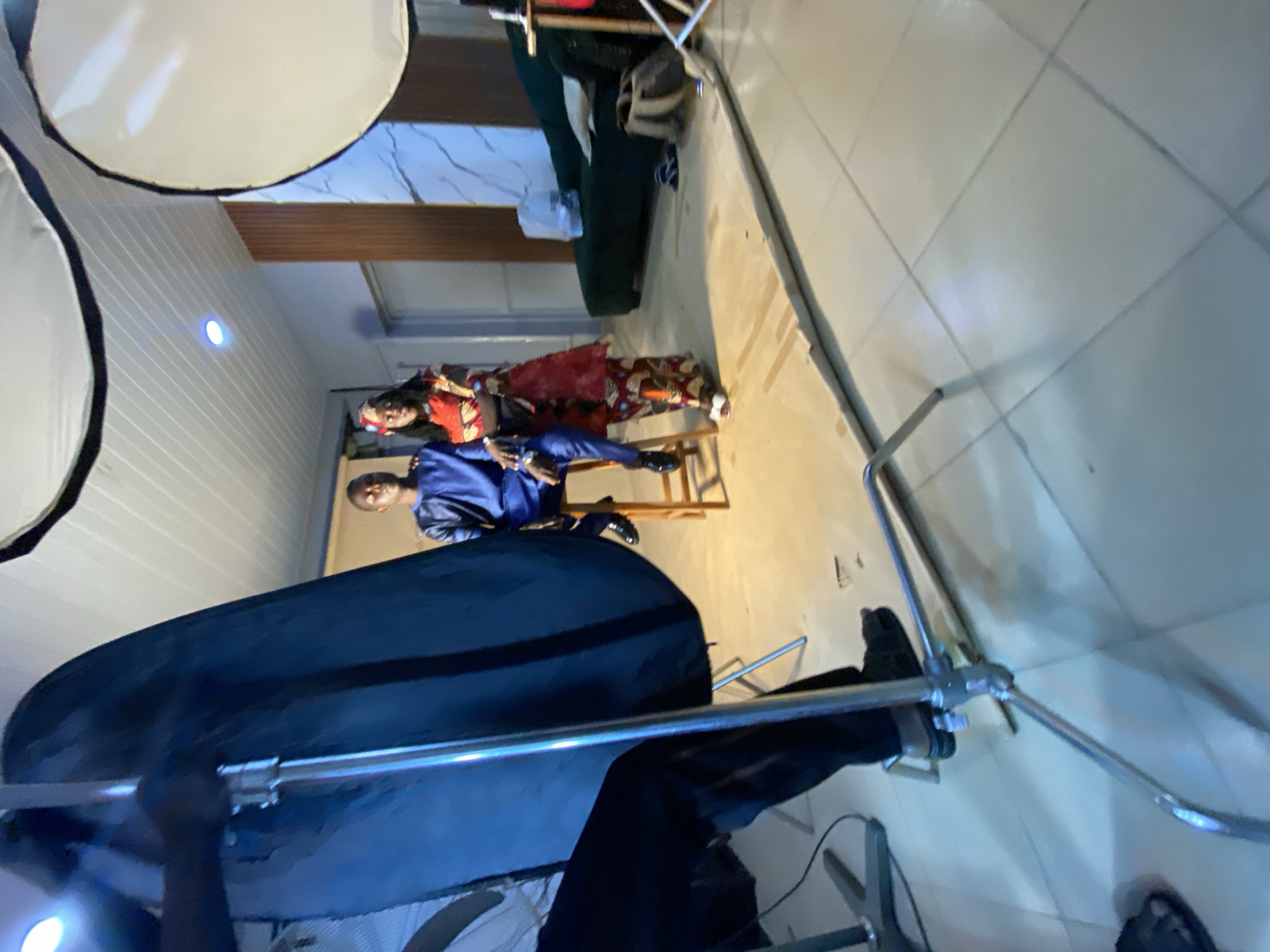 The image captures an indoor scene, possibly a photography studio, in Bukuru, Nigeria. A man and a woman, dressed in what appears to be traditional Nigerian attire, are sitting on a small stool. The man is in a blue outfit, while the woman is in a patterned red and brown outfit. The room is equipped with photography lighting equipment, including softboxes and light stands. There is also a green cushioned bench in the background, set against a wall with what looks like artificial white brick pattern with a brown decorative frame near the top of the wall. The floor is tiled. The lighting and equipment setup suggest a professional or semi-professional photography session.