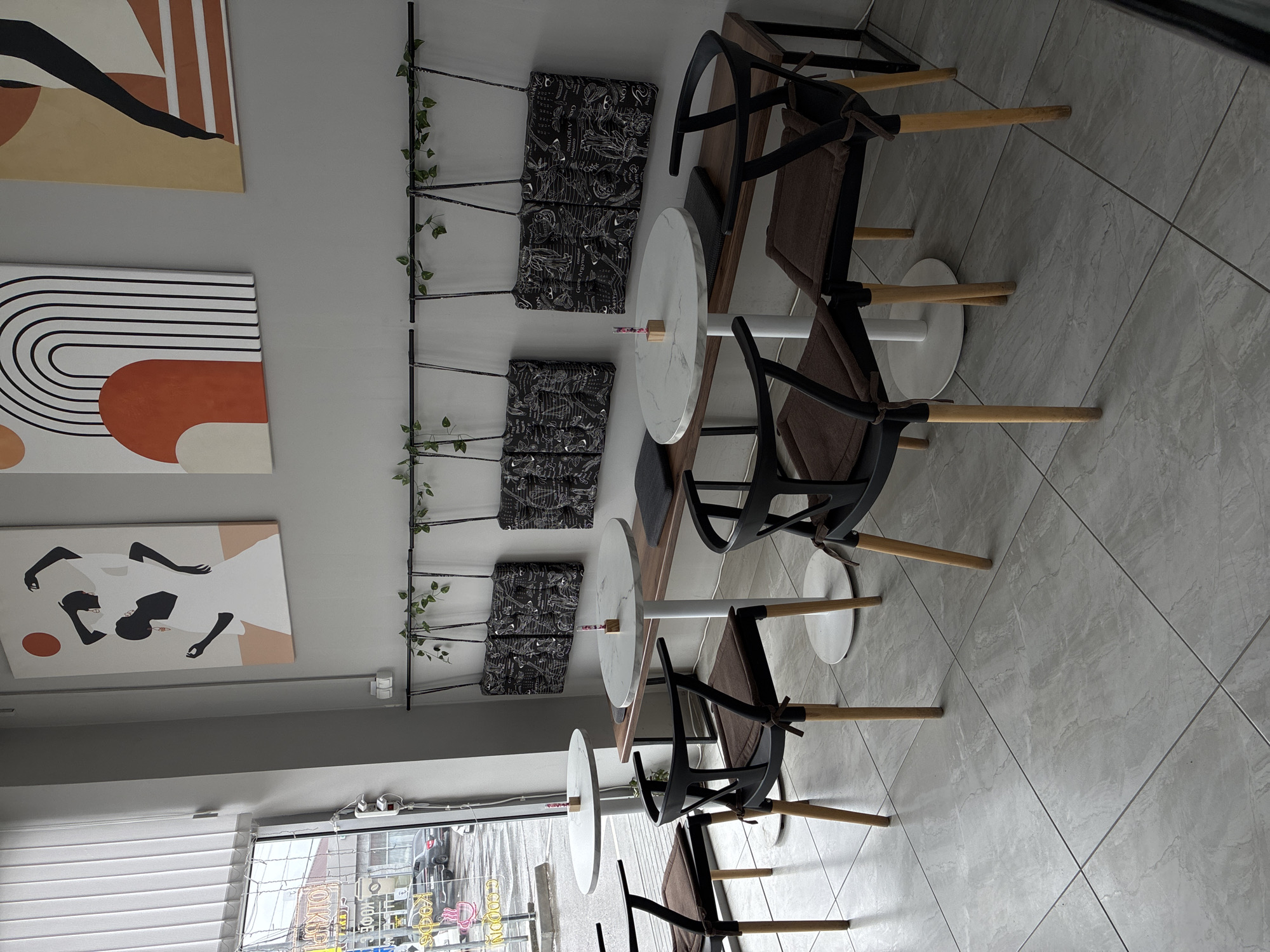 The image shows an interior view of a café, likely in Mozdok, Russia. The scene is composed of several round, white-topped tables arranged along a wall. Each table is paired with two or three chairs featuring black frames and brown cushions. Decorative artwork hangs on the wall, and a plant trellis runs vertically along the wall. The floor is tiled with a light grey material. Outside the window, there are reflections of street signs.