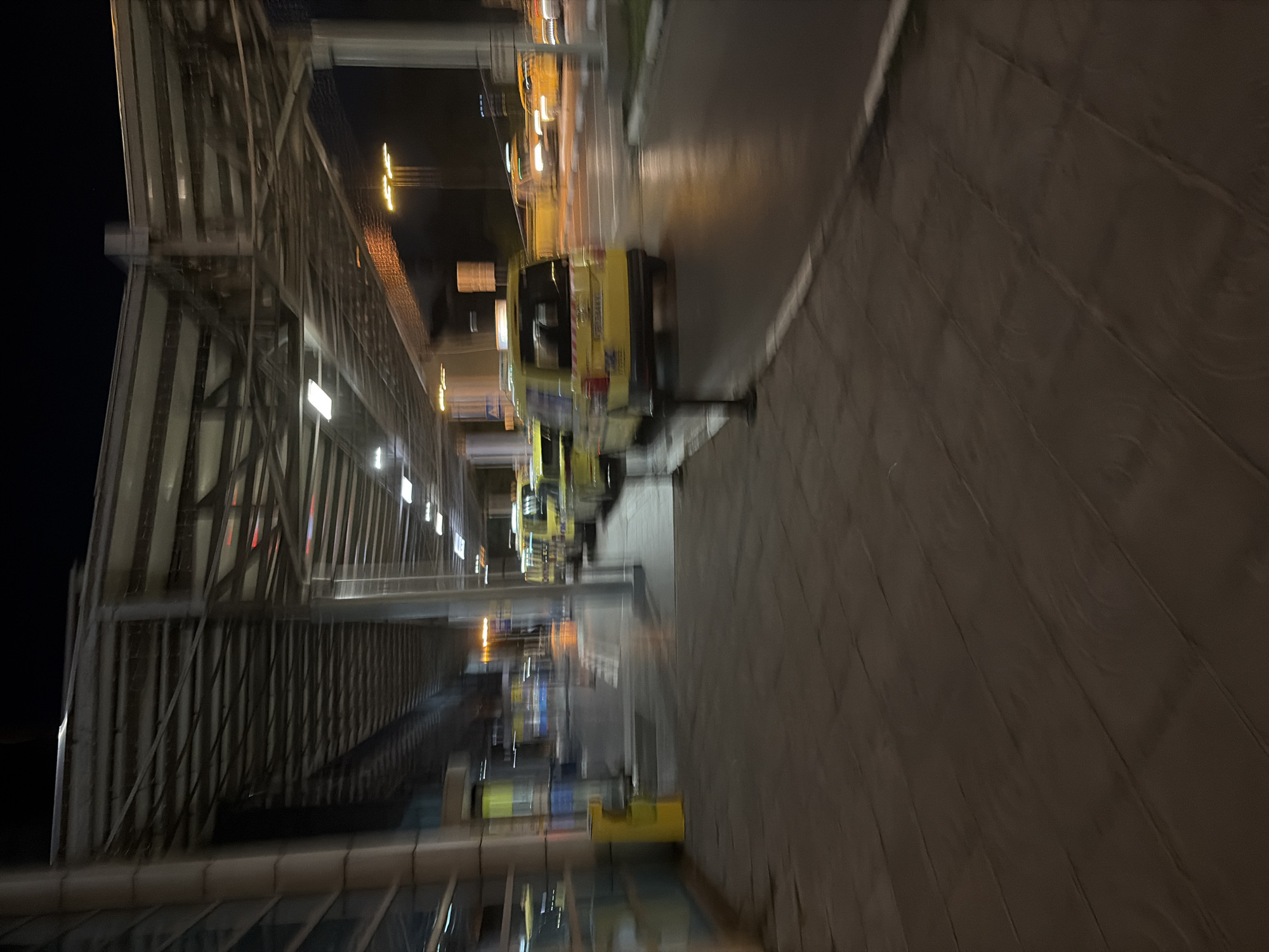 The image depicts a taxi stand at night, likely at an airport or transportation hub, in Sofia, Bulgaria. A row of yellow taxis, characteristic of Sofia's taxi fleet, are lined up on the wet asphalt. The foreground shows a tiled pedestrian walkway, and the background features a covered walkway structure with lighting. The overall scene is dimly lit, with motion blur indicating movement, possibly from vehicles or the photographer.