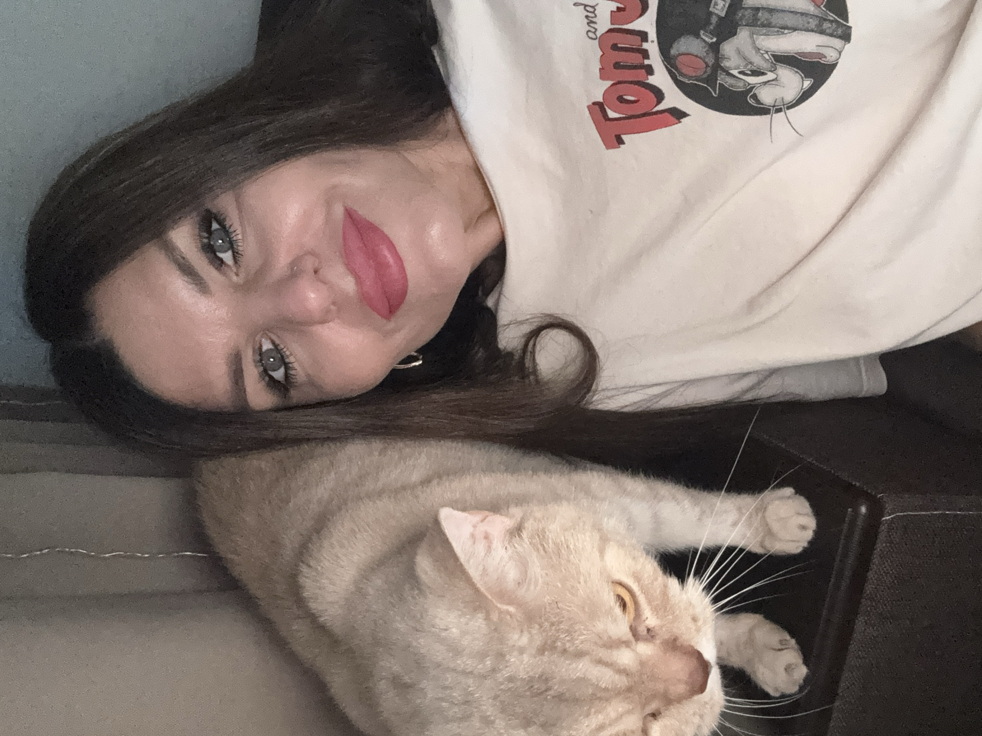 A woman with dark hair and fair skin is taking a selfie. She is wearing a light-colored t-shirt with a graphic of Tom and Jerry. Her lips are painted a bright pink. To her left, a light brown cat is looking down. The background is indistinct, but suggests an indoor setting, possibly a home. There are no identifiable landmarks or features to suggest a specific location within Chaikovsky, Russia.