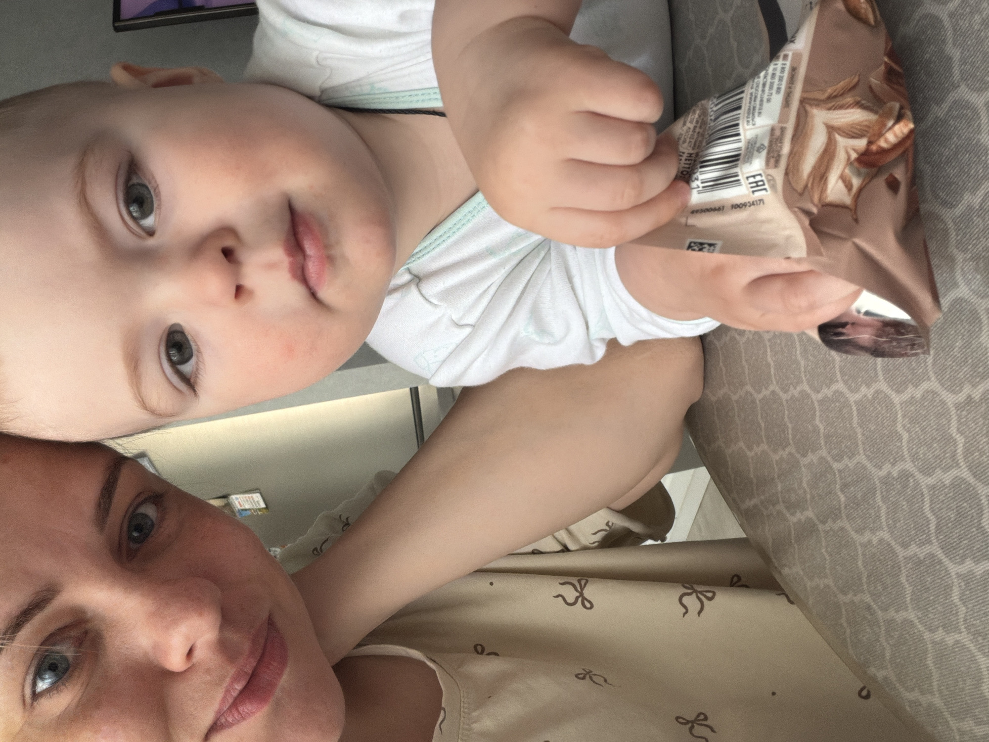 The image features a close-up selfie of an adult woman and a baby, likely taken indoors. The woman's face occupies the lower portion of the frame, angled slightly to her left, with blue eyes looking towards the viewer and a neutral to slight smile. Her light-brown hair is pulled back, and she wears a light-colored top with a pattern of dark bows.
Above and slightly behind her, a baby with light hair and blue eyes looks directly at the camera, displaying a calm or curious expression. The baby is wearing a white garment with light blue trim around the neck and sleeves. In its right hand, the baby holds a brown and beige snack package or bag.
Notable details on the package include an "EAC" (Eurasian Conformity) logo, a barcode, and numerical sequences "49300661" and "100934171". Other text in Cyrillic script and potentially English is partially visible but largely unreadable due to the angle and focus. The background consists of a light wall or surface behind the woman's head, and a patterned gray fabric (likely upholstery) to the right. The overall impression is a casual, intimate moment captured between the two subjects. The setting appears to be a home in Votkinsk, Russia, based on provided context.
