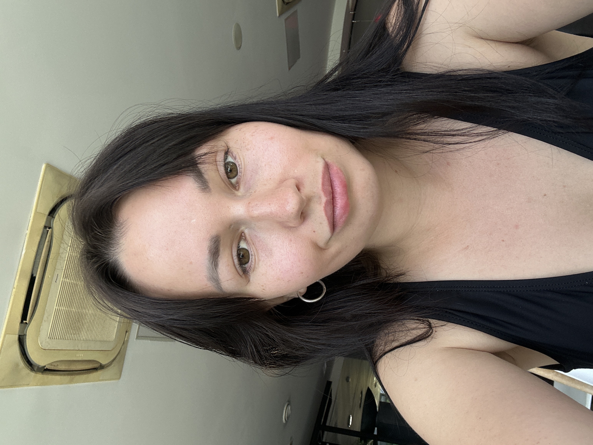 A medium-shot, chest-up selfie of a woman with long, dark hair and brown eyes is captured. She is wearing a black swimsuit and a silver hoop earring. The woman has a light skin tone with freckles across her nose and cheeks. Her hair is partially covering her face and shoulders. The background consists of a white ceiling with a beige air conditioning unit and a glimpse of what appears to be an indoor space with ceiling lights. The lighting suggests an indoor setting, possibly during the day. There is no discernible connection to Saint Petersburg, Russia, based on the provided visual information.