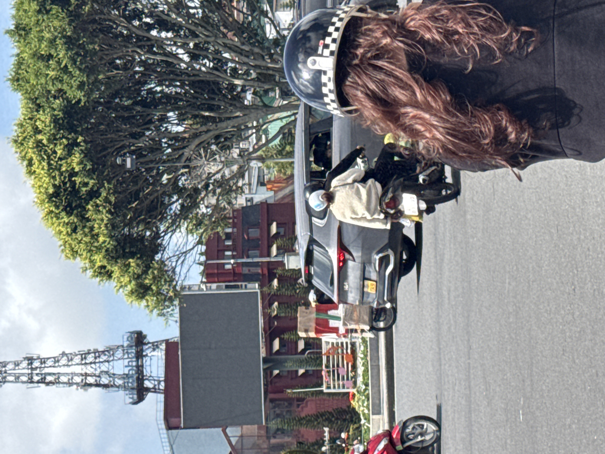 In Da Lat, Vietnam, on a clear day, a woman wearing a black jacket and a black and white checkered helmet rides on the back of a motorcycle, with her long, wavy, reddish-brown hair flowing. In front of her, two individuals are riding a motorcycle, with the passenger wearing a light-colored jacket and a white helmet. They are passing a gray Kia Seltos SUV, which has a yellow license plate. To the left, a red motorcycle is parked. In the background, a large digital billboard is visible, along with a tall radio tower bearing the "Viettel" logo. Further back, red buildings with arched windows and trees line the street, suggesting an urban environment. The scene appears to be a typical street view in Da Lat, likely during daylight hours.
