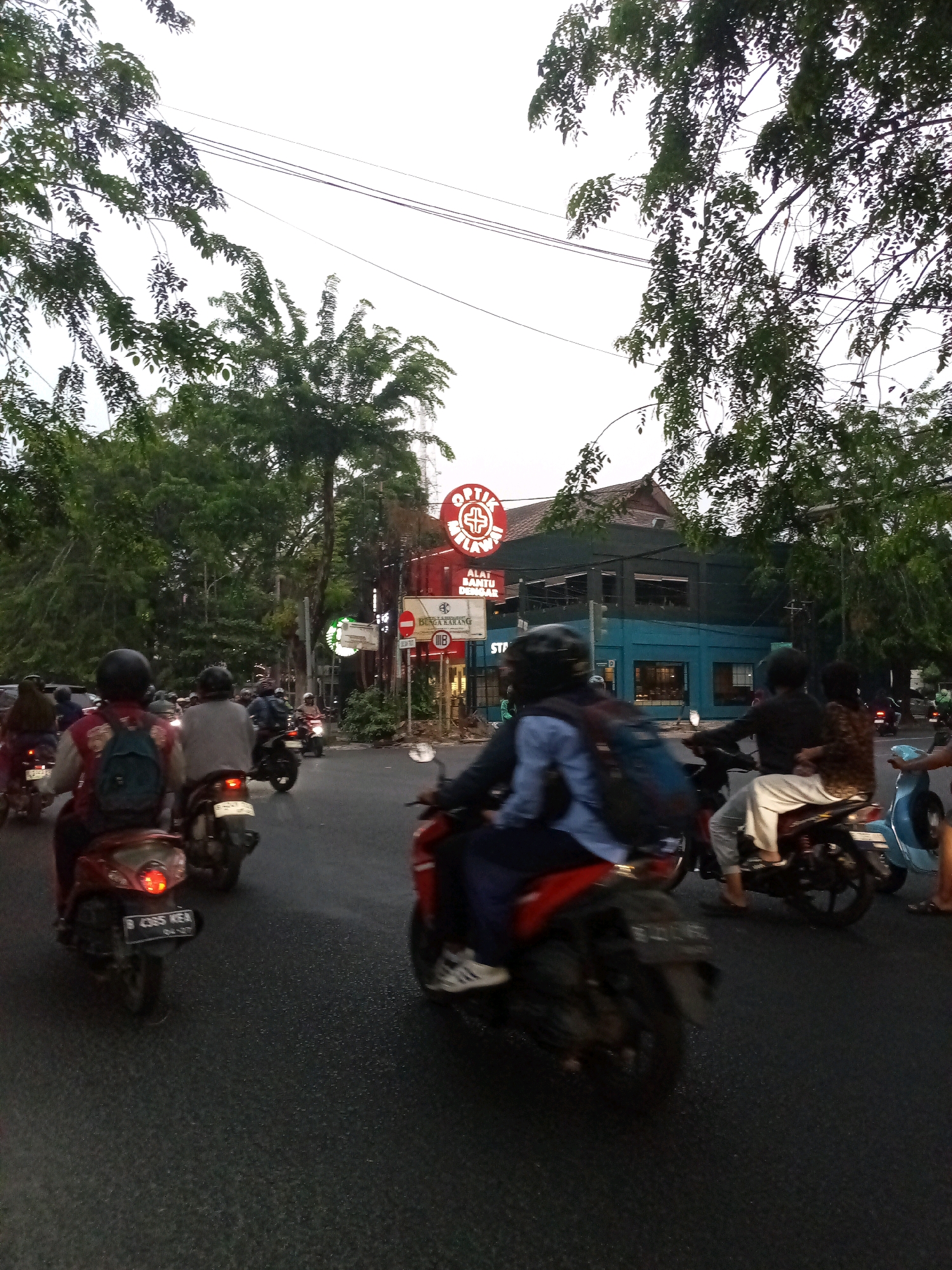 The image depicts a street scene in Bekasi, Indonesia, likely in the evening, as indicated by the overcast sky and artificial lighting. A procession of motorcycles and scooters navigates a paved road lined with trees. On the right side of the road, a building with a blue facade features a prominent red circular sign with a white cross, identifying it as "OPTIK." Below this sign, other signage indicates "ALAT BANTU DENGAR" (hearing aids) and "STATIONERY." Further down, a blue and white sign for "BUNGA KARANG" is visible. Several individuals are riding motorcycles, some with backpacks, and one person is on a light blue scooter. The road surface shows signs of being wet or reflective. Power lines are visible overhead, crisscrossing through the tree canopy.
