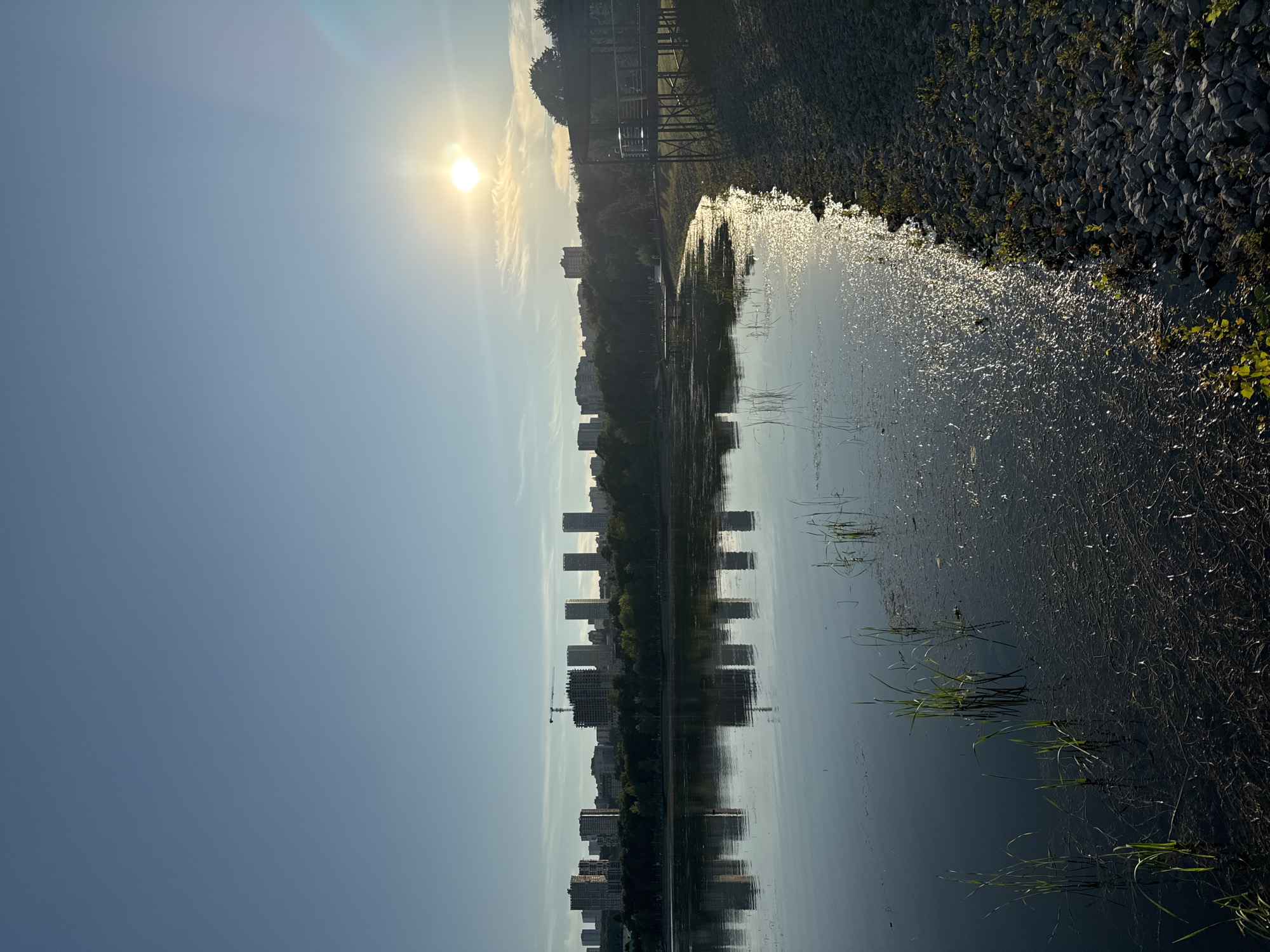 The image depicts a wide, calm body of water, likely a lake or reservoir, in Ufa, Russia. On the far side of the water, a dense line of trees forms a natural barrier, behind which a cluster of modern high-rise buildings and construction sites, including a prominent crane, punctuate the skyline. The sun, positioned in the upper right quadrant of the image, casts a bright, warm glow across the scene, reflecting off the water's surface and creating a shimmering effect. The water's edge on the right side of the image is lined with stones and a small wooden structure, possibly a gazebo or viewing platform, suggesting a park or recreational area. The overall atmosphere is serene and hints at an urban landscape integrated with nature.