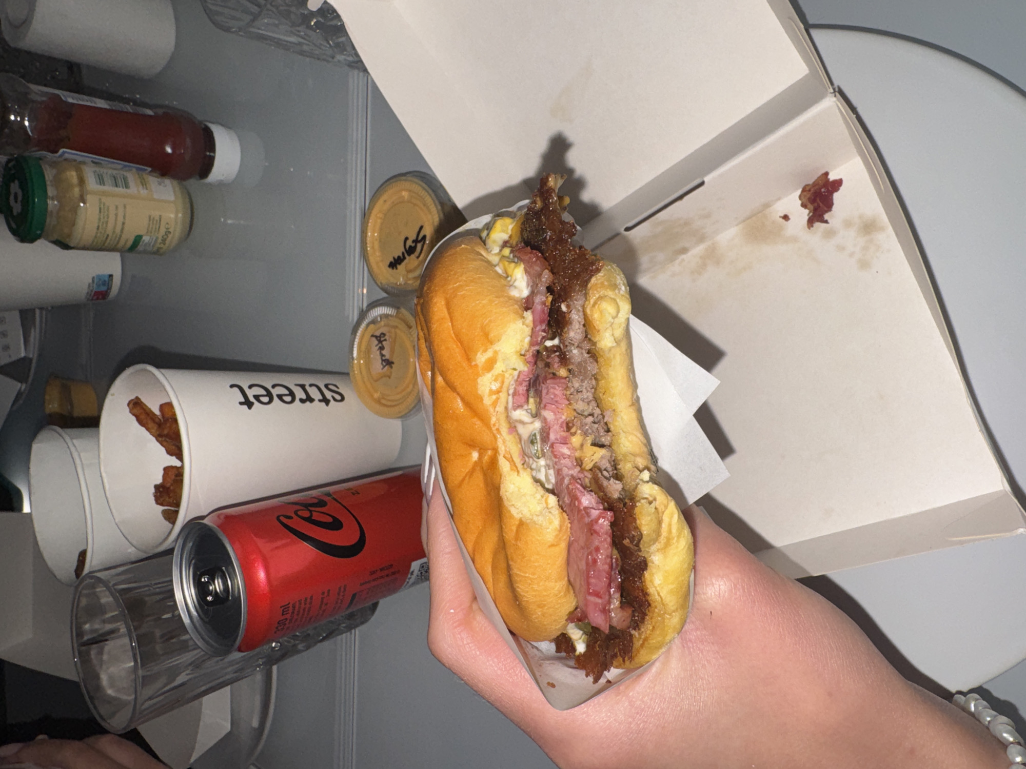 A partially consumed burger is held by a hand, positioned within an open food container. Adjacent to the container are two small condiment cups. The burger appears to have a beef patty, relish, and other toppings inside a soft bun. A Coca-Cola can is located nearby. Further behind is a container of fries and a condiment bottle. The surroundings appear to be inside a refrigerator. The word "street" is visible on one of the containers. The scene is likely in Milan, Italy, indicated by "Street" brand of burger.