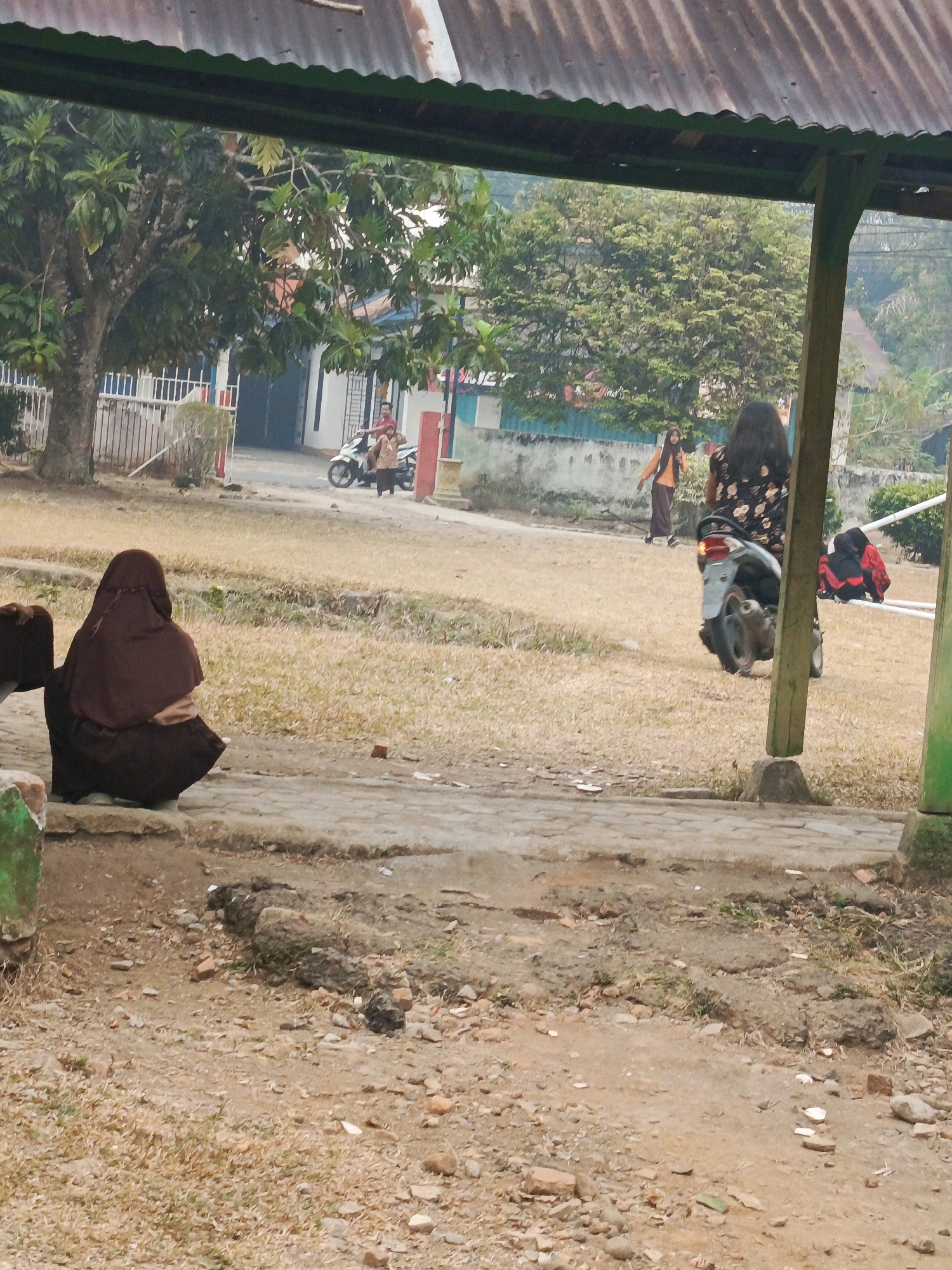 The image depicts an outdoor scene, likely in a public area or schoolyard, with dry grass and a paved walkway. In the foreground, a person wearing a brown hijab and dark clothing is seated facing away from the camera, looking towards the center of the frame. To the right, a motorcycle is parked, and a person with long dark hair wearing a floral-patterned dress is standing near it. In the background, another person in an orange top and long skirt is walking, and a man is seated on a motorcycle. There are buildings and trees visible further in the distance, suggesting an urban or semi-urban environment. The presence of a metal roof structure with corrugated iron panels partially obscures the top of the image, indicating a covered outdoor space. The overall impression is one of a casual gathering or moment in daily life.