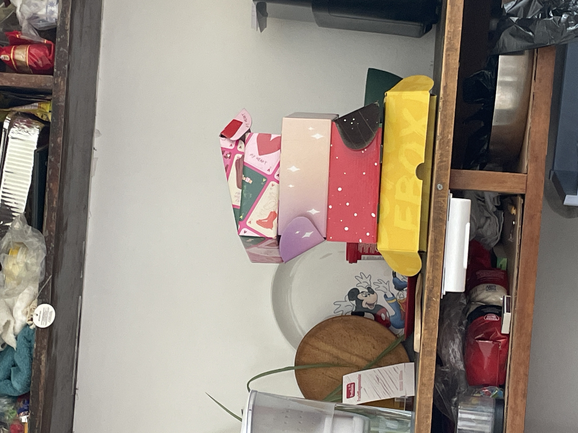 The image shows a cluttered shelf or cupboard, possibly located in Plovdiv, Bulgaria. On the left, a wooden structure holds an assortment of items, including food packaging, plastic bags, and a cleaning cloth. To the right, another wooden shelf unit displays gift boxes of varying sizes and colors, a plate with a Mickey Mouse design, a round object that appears to be a cake base, and additional containers or packets. The background consists of a plain white wall. The overall impression is one of disorganization and haphazard storage.