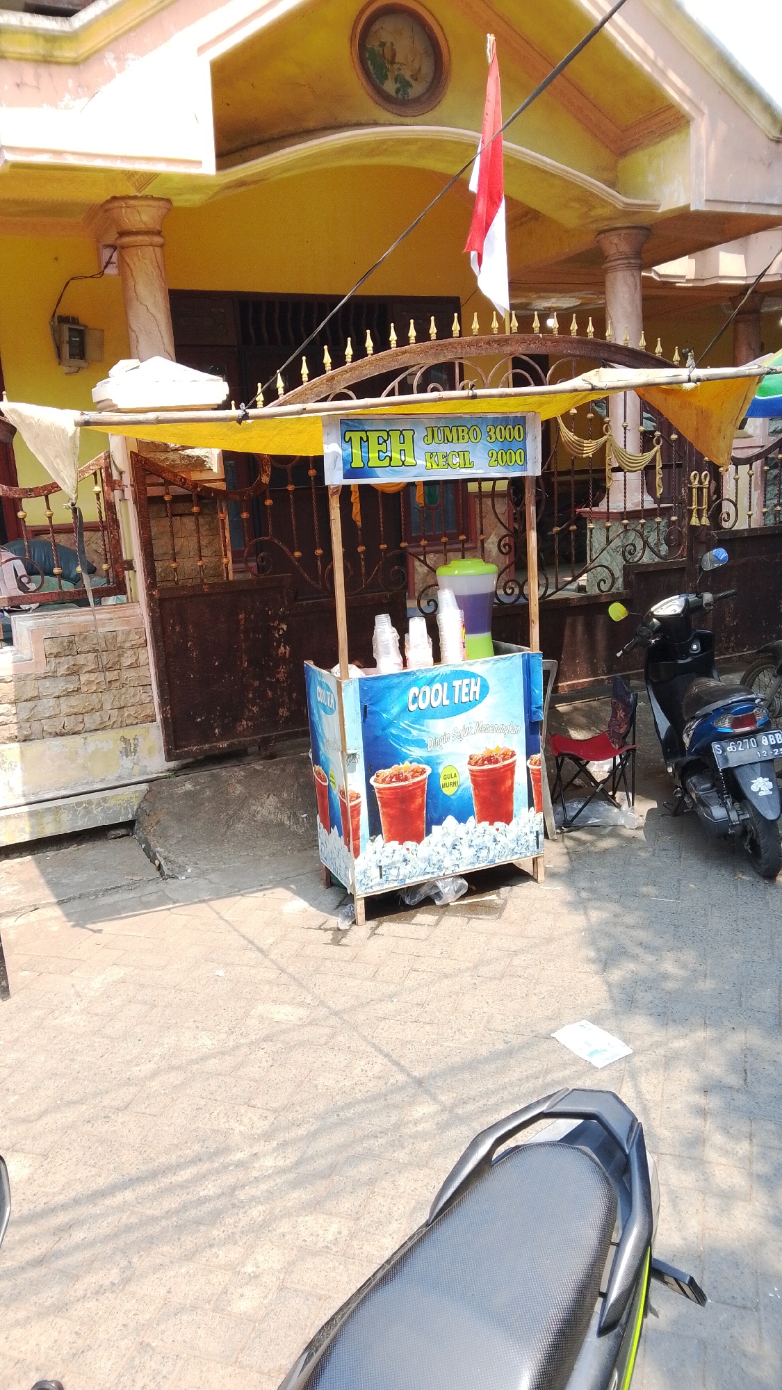 In the city of Mancar, Indonesia, an outdoor beverage stall is set up on a paved area. The stall features a yellow canopy and a display cart with "COOL TEH" branding and images of iced tea. Several plastic cups are visible on top of the cart. A large beverage dispenser is also present. A motorcycle is parked to the right of the stall. In the background, a house with a yellow facade and a decorative metal gate is visible. An Indonesian flag, red and white, is flying near the house. The ground is a mix of concrete and paving stones.