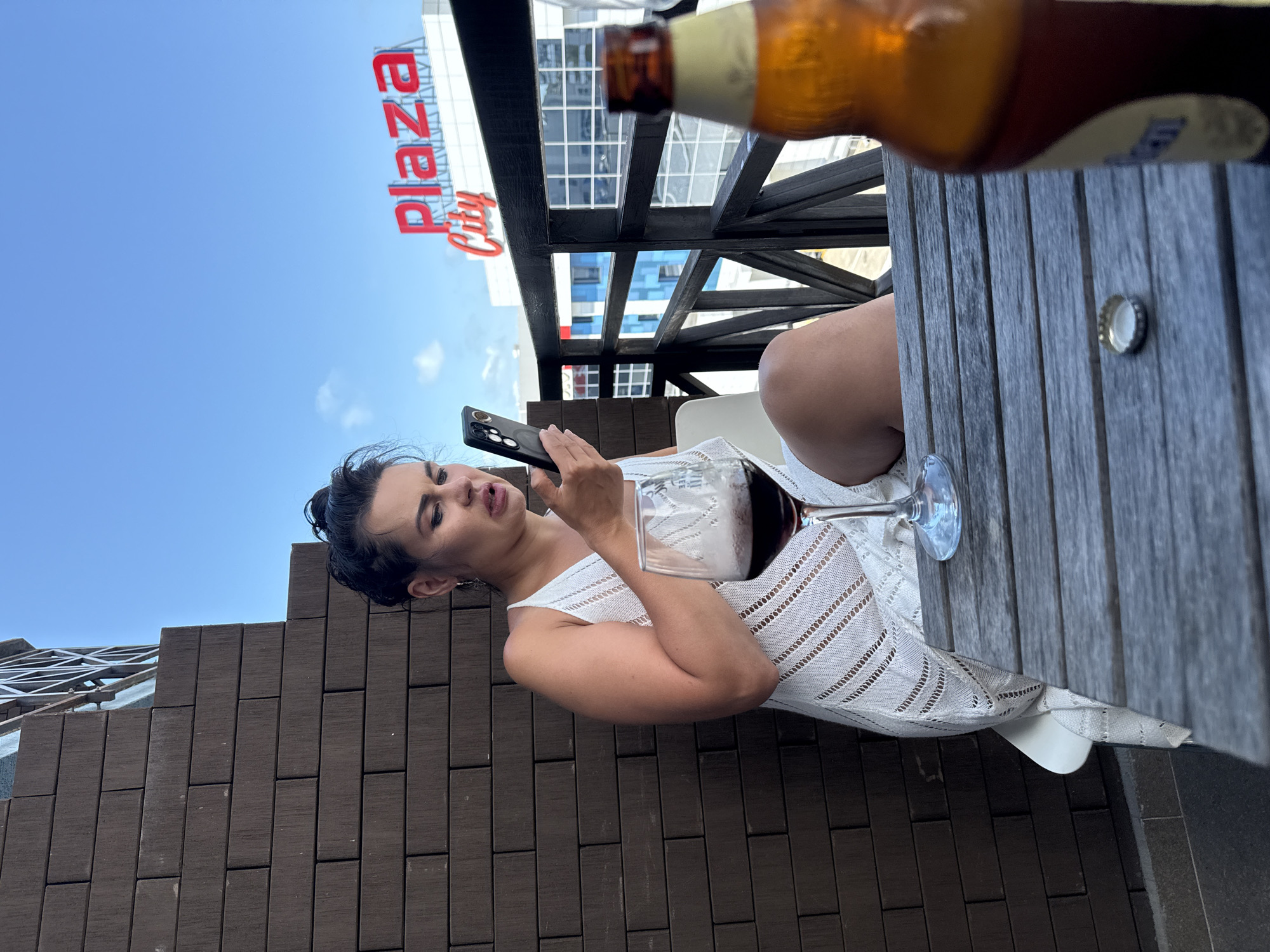 A woman is sitting at an outdoor table, holding a smartphone. She is wearing a white, crocheted dress. To her right, a glass of dark red liquid, likely wine, is on the table. A beer bottle is partially visible in the foreground, and a bottle cap rests on the wooden table. The background shows a building with a large red sign that reads "Plaza City." The sky is blue with some scattered clouds. The scene appears to be set during the daytime. The woman's expression suggests she is engaged with her phone. The setting is an outdoor seating area, possibly a restaurant or bar, with wooden decking and a dark wooden railing. The view includes a city skyline, hinting at an urban location.