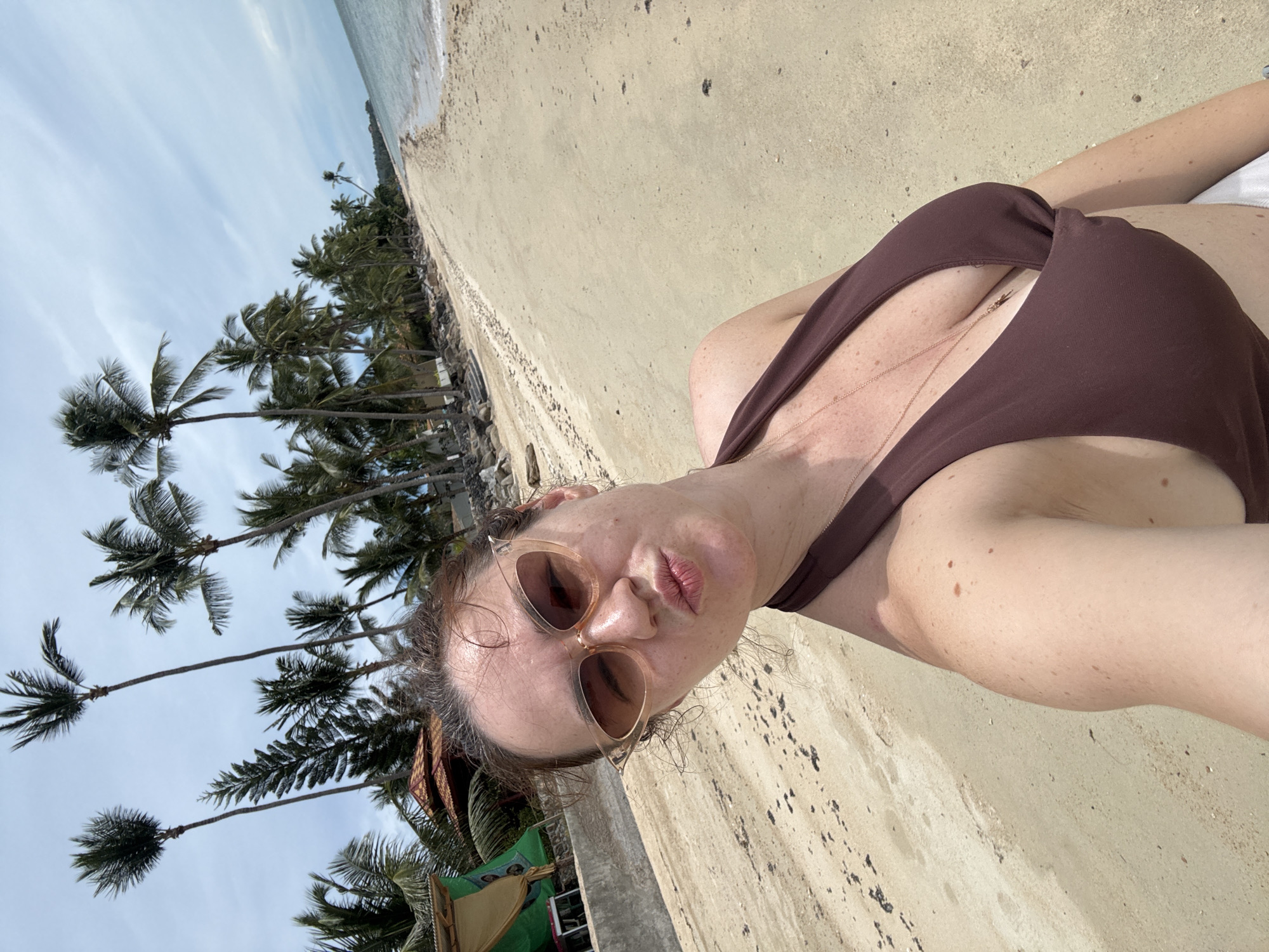 A woman stands on a sandy beach in Baan Tai, Thailand, taking a selfie. She is wearing a brown bikini and large sunglasses. The beach is lined with palm trees, and the ocean is visible in the background under a cloudy sky.