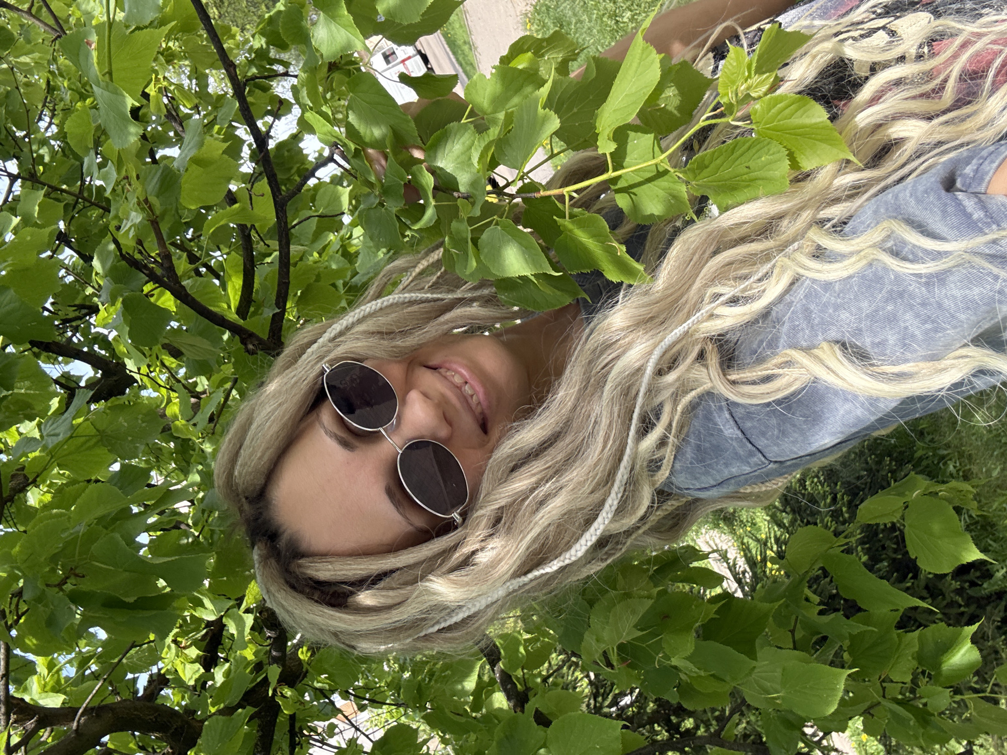 A young woman with blonde hair and sunglasses is pictured from a low angle. She is surrounded by green leaves and branches. She is smiling. The location is Rusanovo, Russia.
